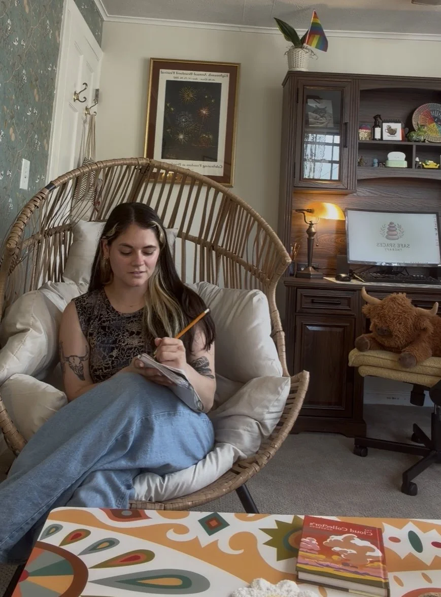 A young woman with tattoos on her arms sitting in a wicker chair in a cozy room, writing in a notebook with a pencil, surrounded by pillows. Behind her, there's a framed picture of fireworks, a wooden cabinet with decorative items, and a desk with a mouse and monitor displaying a spa logo. A plush teddy bear is on a chair next to the desk, and a rainbow party hat is on top of a white basket.