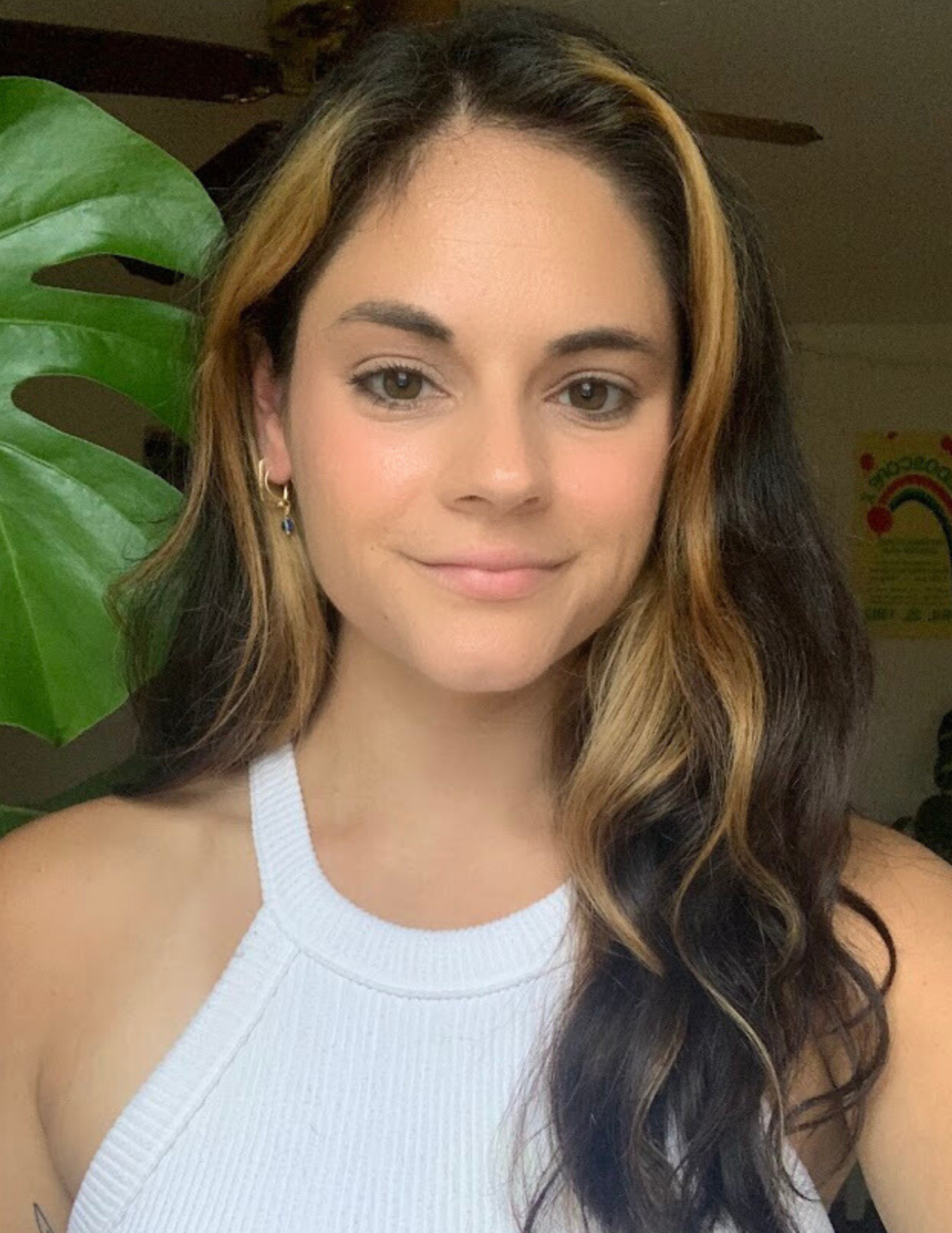 A young woman with long, wavy hair that is dark at the roots and blonde at the tips, wearing a white sleeveless top, standing next to a large green plant, smiling softly at the camera.