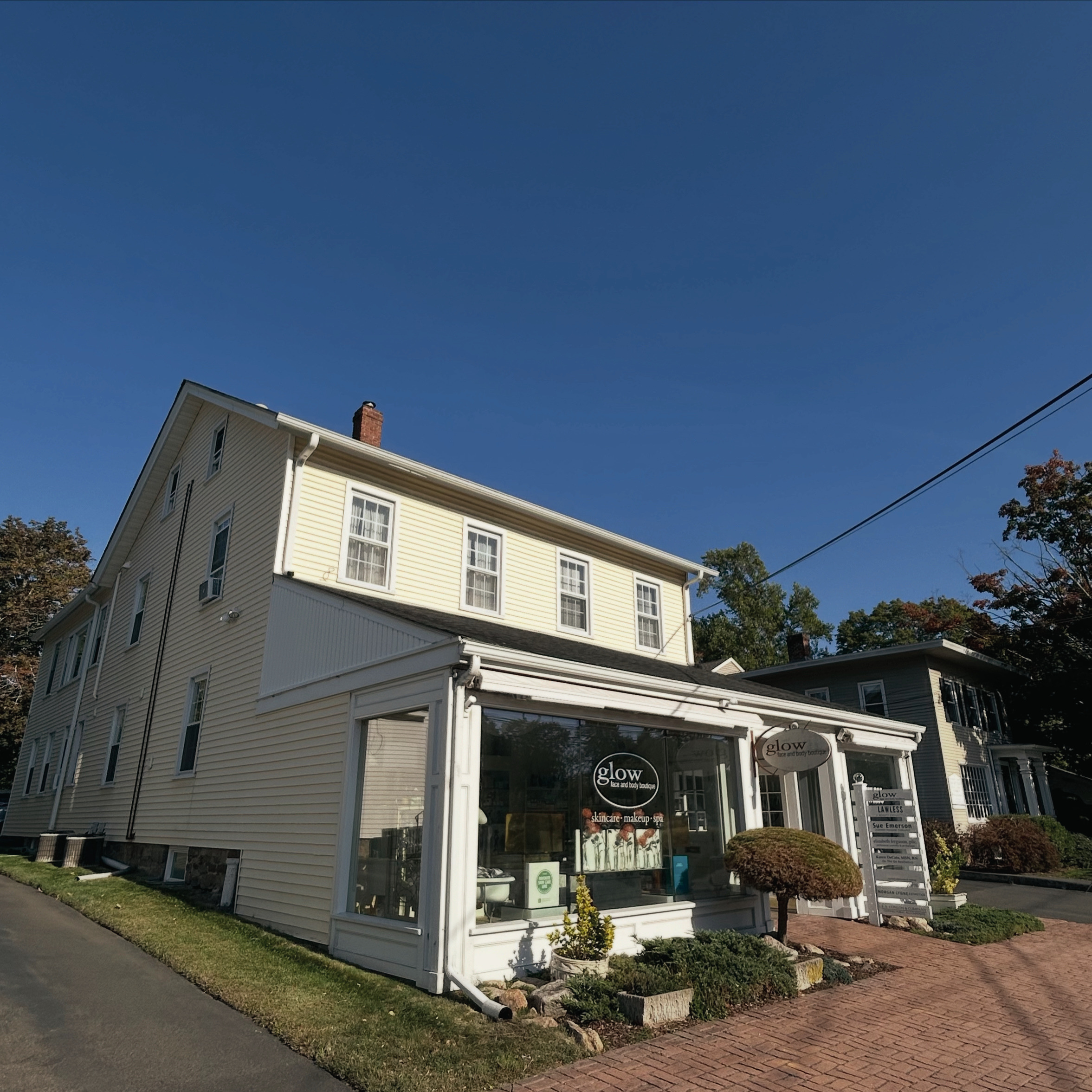 A corner store with a large glass window and a sign that says 'glow' in a small shopping complex. The store is adjacent to a yellow multi-story house with white trim and multiple windows, set against a background of green trees and a clear blue sky. Paved walkway and small landscaped area with plants and trees are in the foreground.