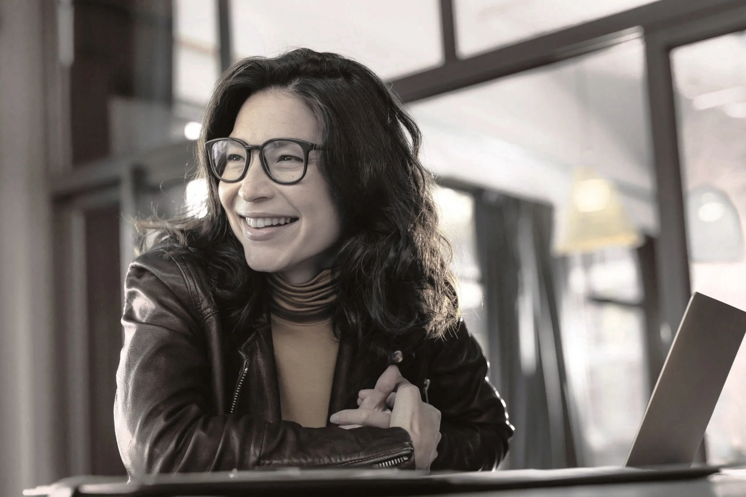 Eine lachende Frau mit schwarzen Haaren, Brille und dunkler Lederjacke sitzt an einem Tisc. Vor ihr liegt ein Laptop in einem modern eingerichteten Raum.