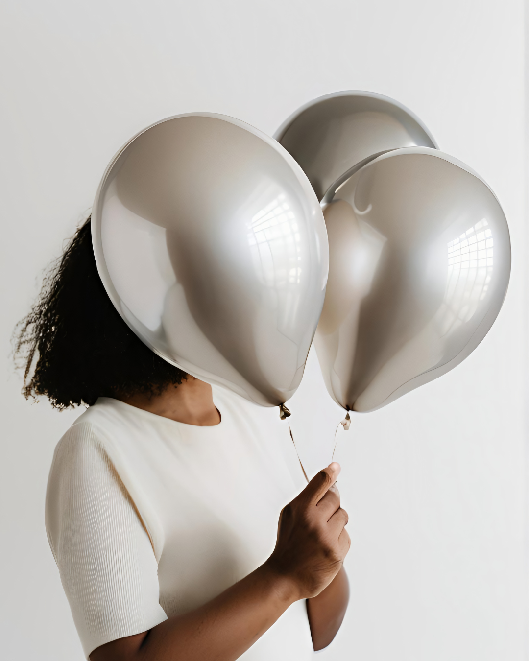 Frau mit schwarzen Haaren und weißem Pulli hält drei silberne Luftballons vor das Gesicht.