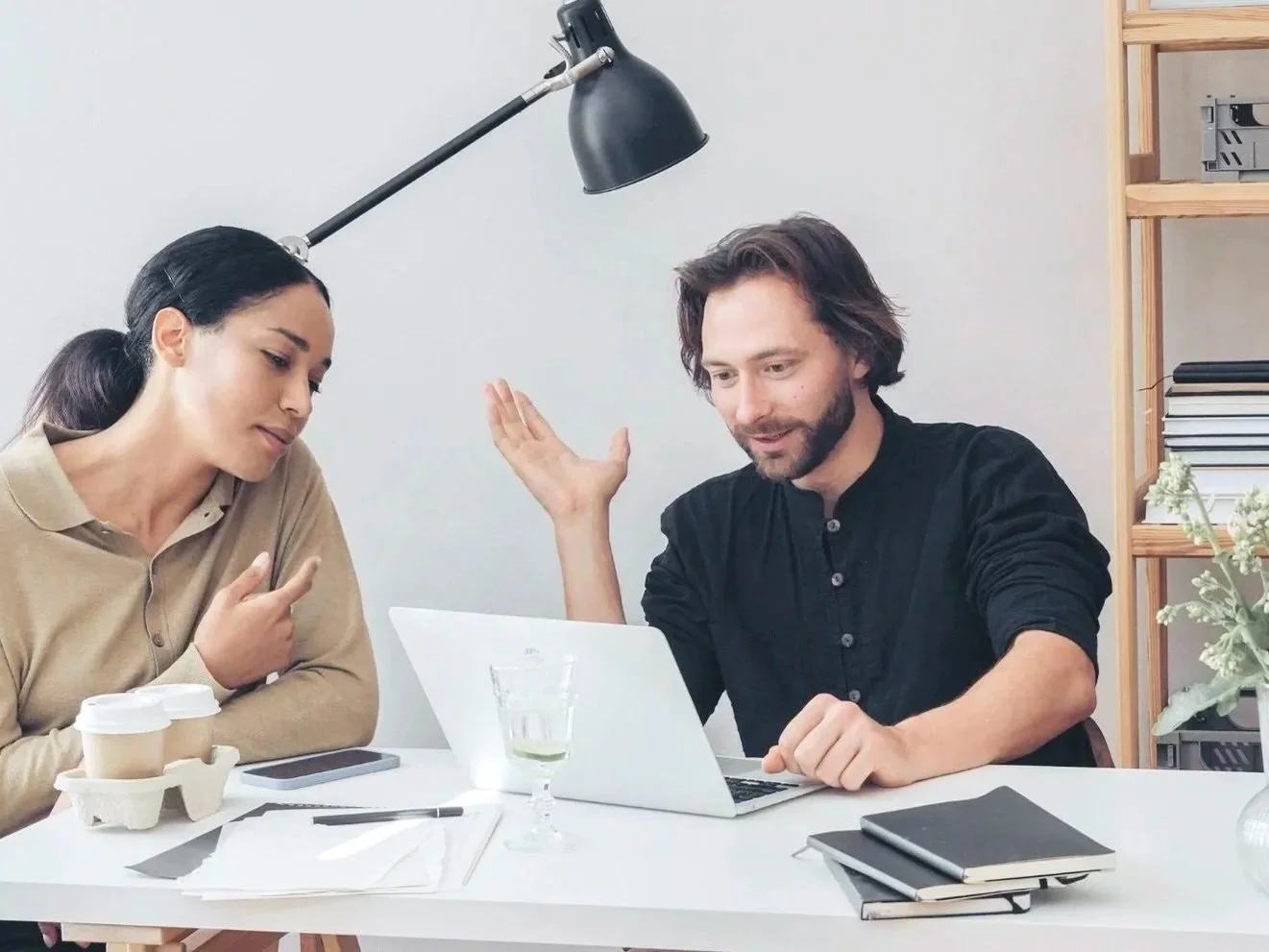 Zwei Menschen sitzen an einem weißen Tisch, einer zeigt auf einen Laptop während der andere interessiert zuschaut, im Hintergrund ein Regal mit Pflanzen und Büchern sowie einer großen Stehlampe.