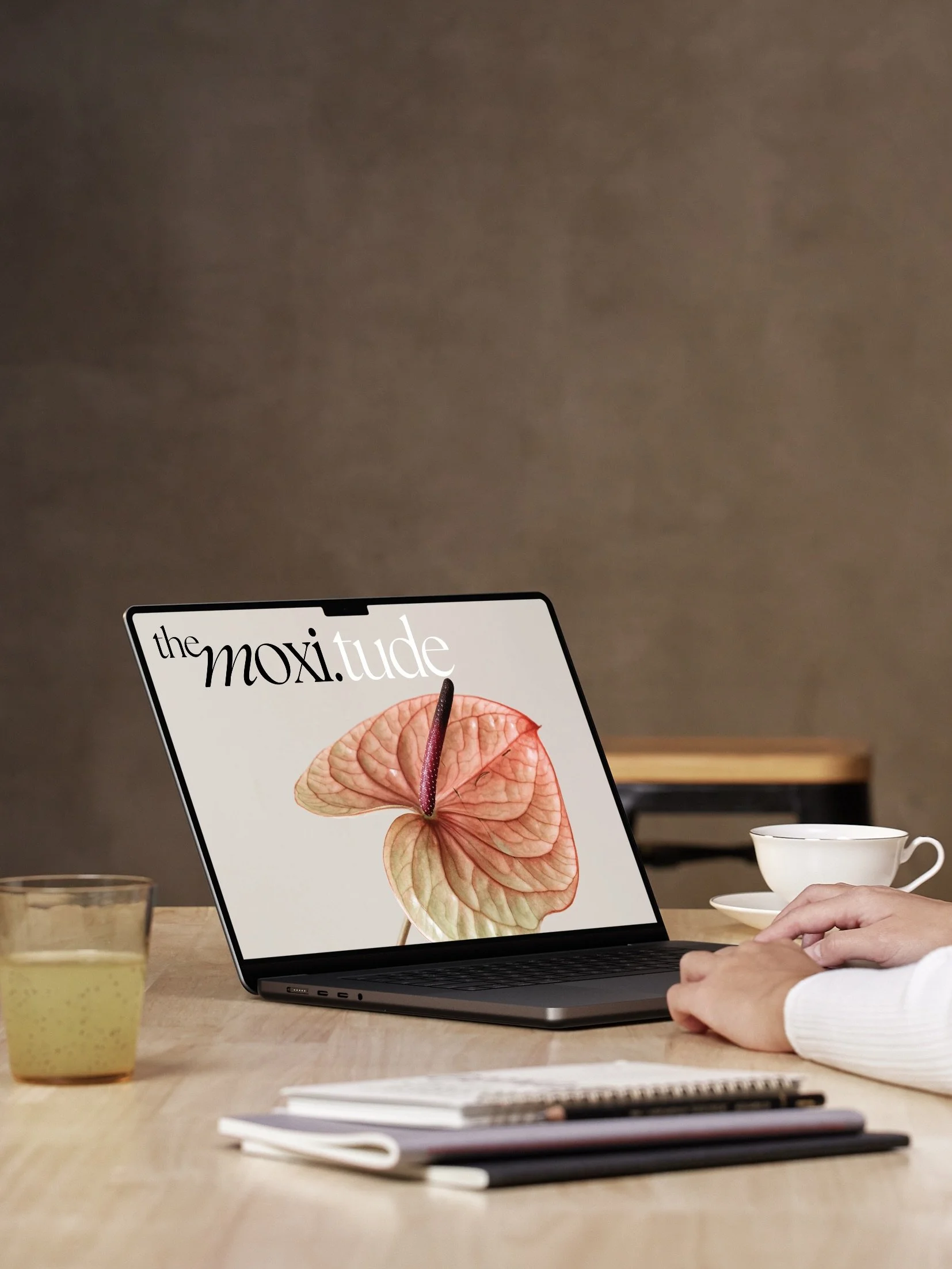 Ein Laptop auf einem Holztisch zeigt das Magazin "The Moxi.tude" mit einer Nahaufnahme mit einer Anthurie auf dem Bildschirm, umgeben von einer Tasse Kaffee, einem Glas Saft, einem Notizbuch und einem Stift.