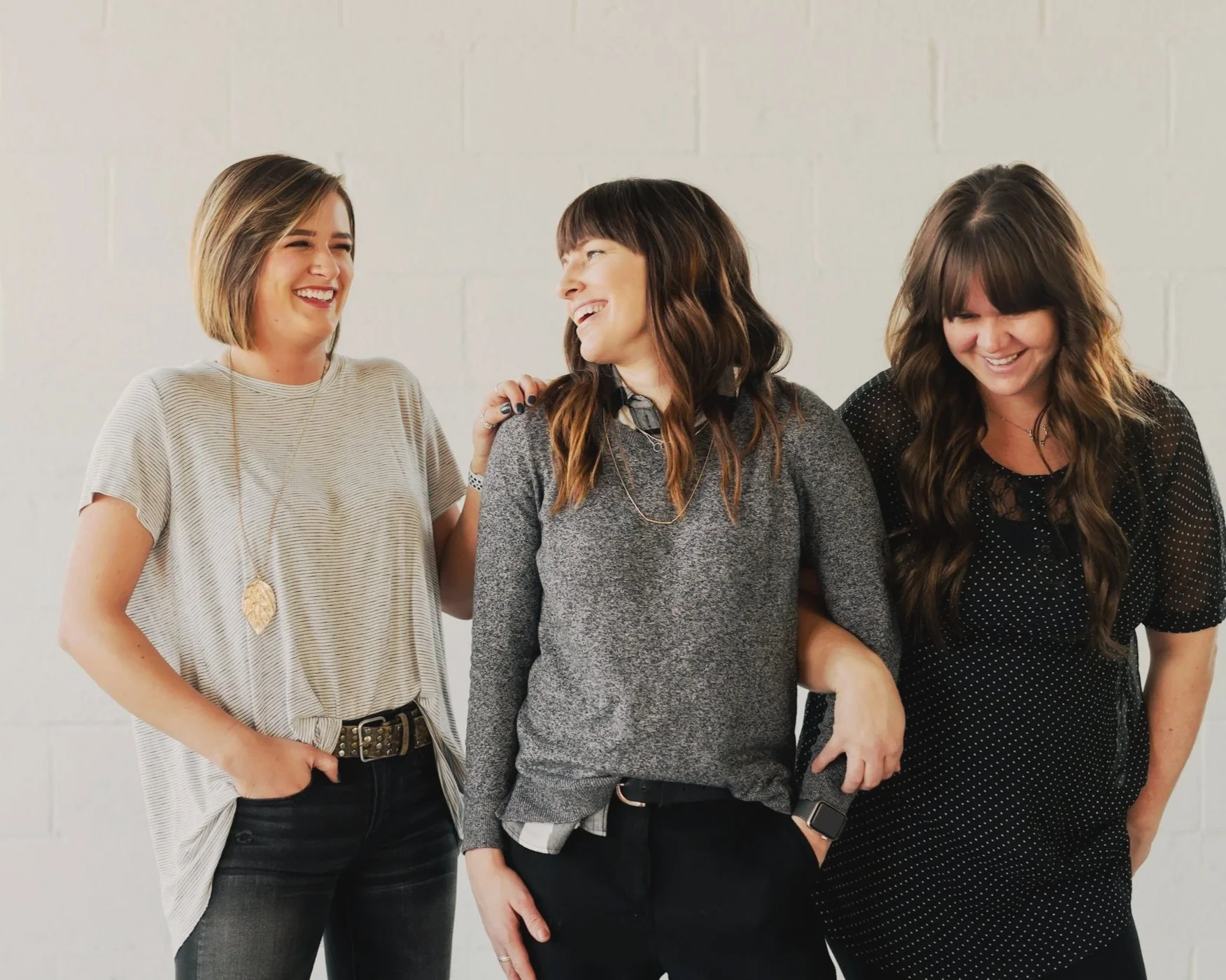 drei Frauen, eine im T-Shirt mit Pagenschnitt, zwei mit dunklen langen Haaren, grußer Pulli und schwarzes T-ShirtFrau im beigen Polo mit Pferdeschwanz sowie Mann mit Bart und schwarzem Hemd am Tisch mit Laptop, dahinter schwarze Lampe