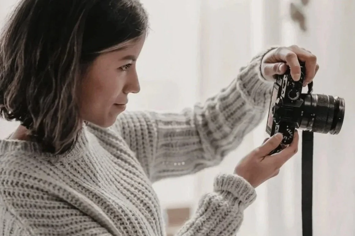Frau in hellgrauem Pullover hält Kamera und schaut durch Sucher.