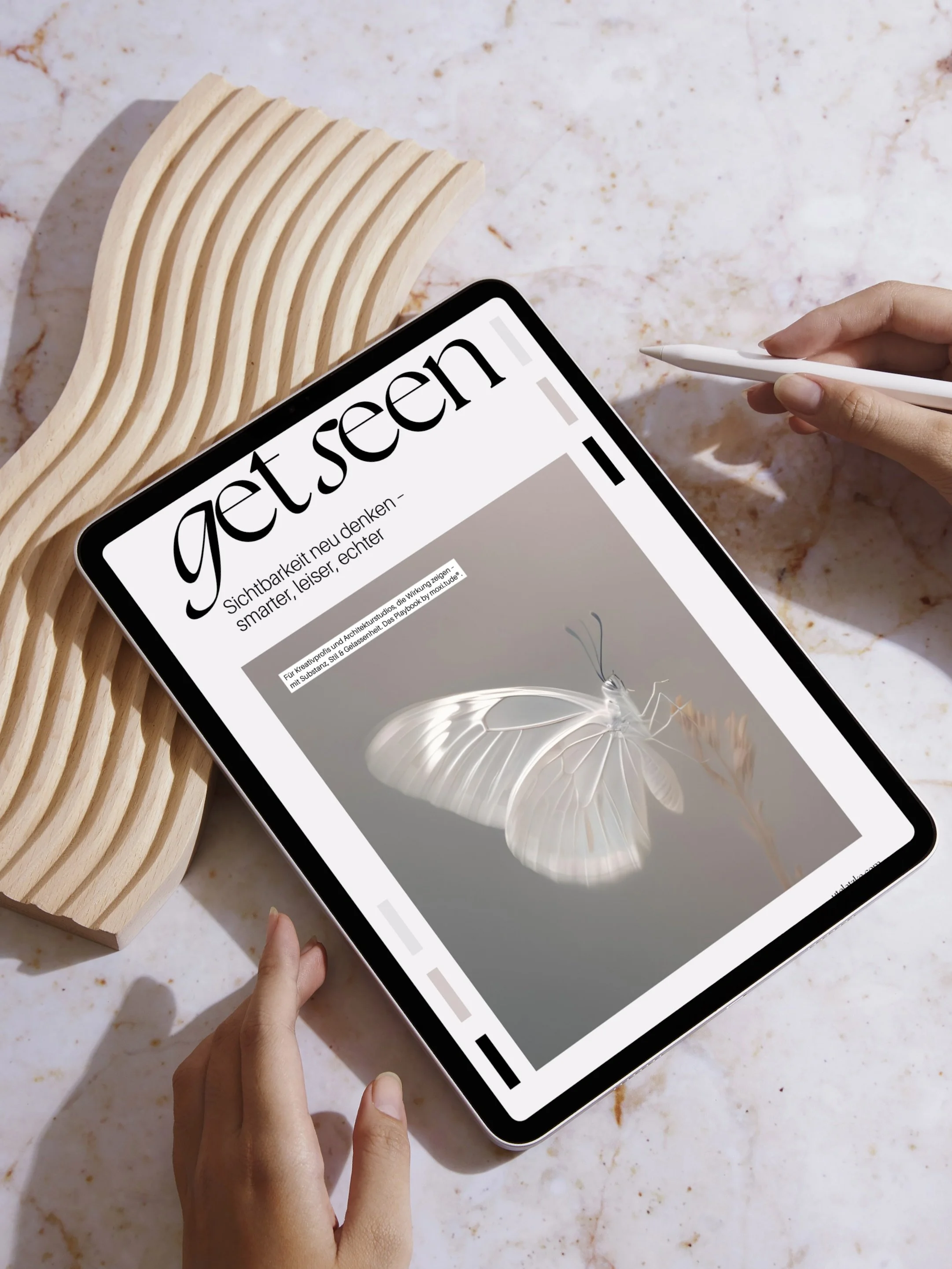 iPad mit Cover von Get Seen mit Schmetterling, liegt auf Tisch und Holzform, eingerahmt von zwei Händen