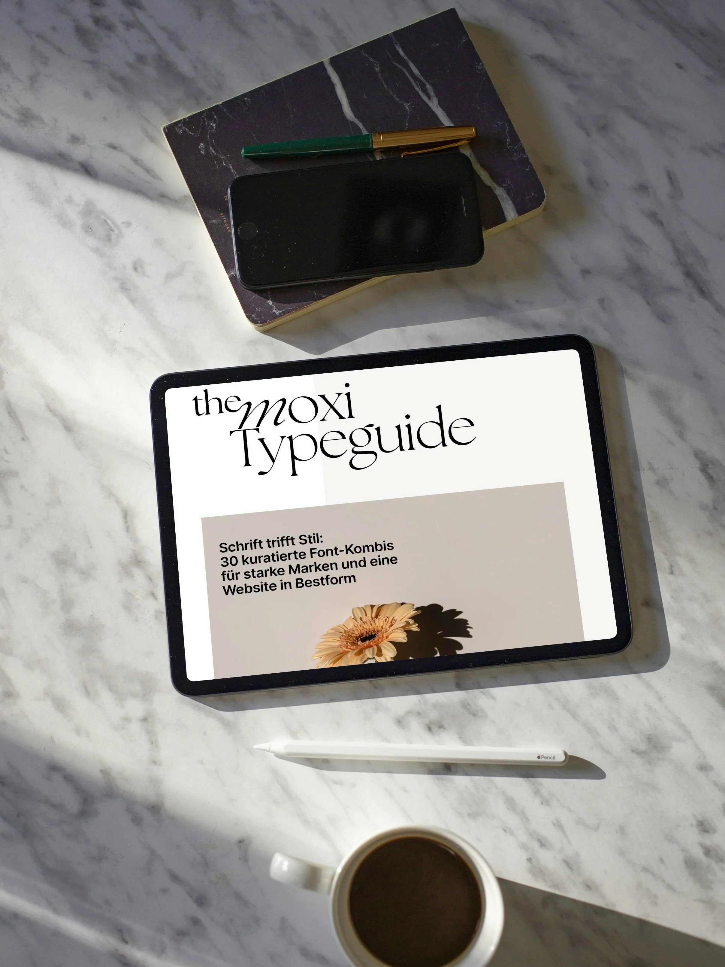 Tablet zeigt ein Typografie-Guide mit dem Titel "the Moxi Typeguide" und einem Untertitel in Deutsch. Ein Notizbuch, ein Smartphone, ein Stift, eine Tasse Kaffee und ein weiterer Stift liegen auf einem weißen Marmortisch.