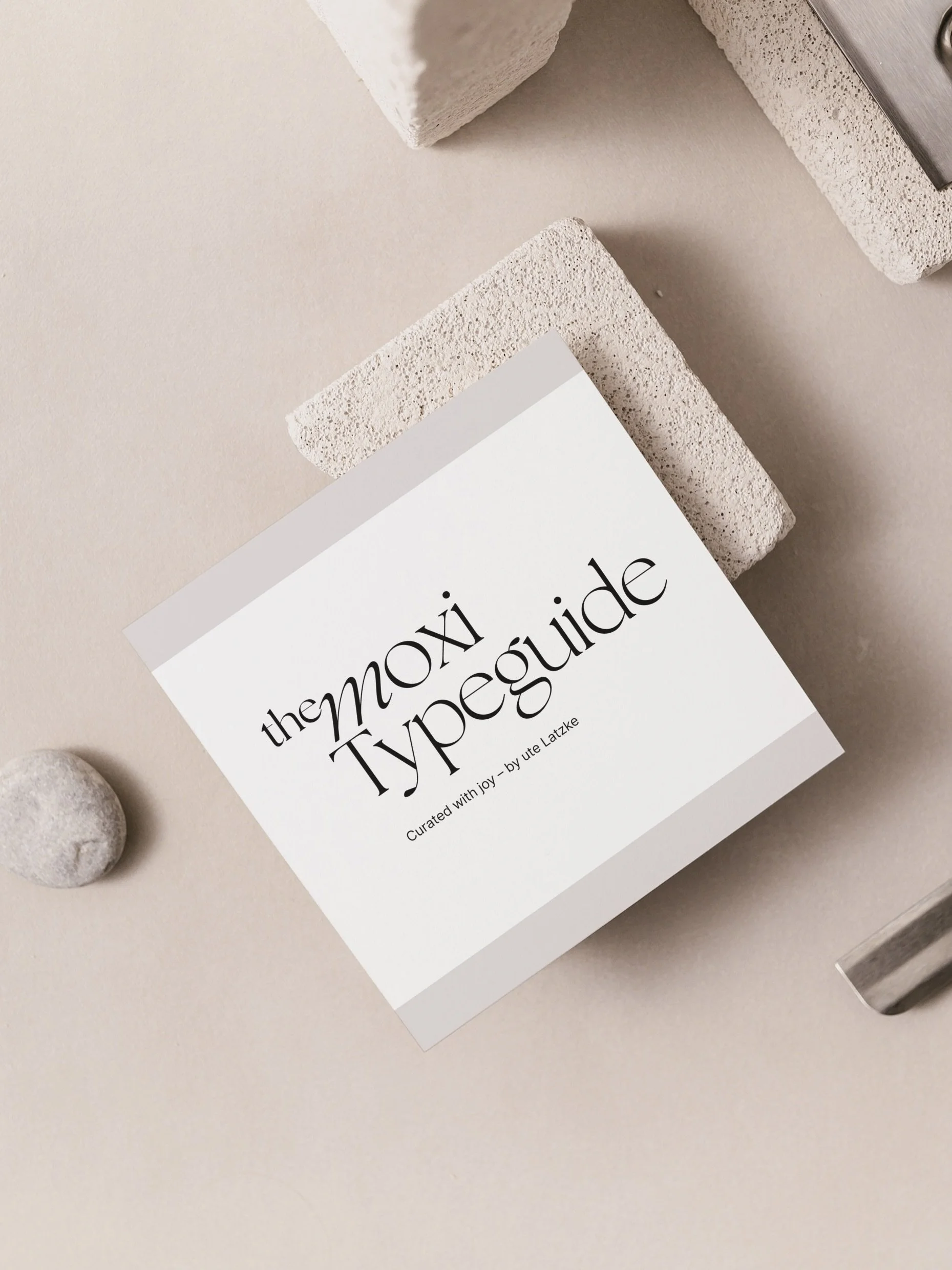 Ein weißes Buch mit schwarzem Text, das den Titel "the moxi Typesguide" trägt, liegt auf einem beige Untergrund, umgeben von Steinen und rechteckigen Bausteinen.