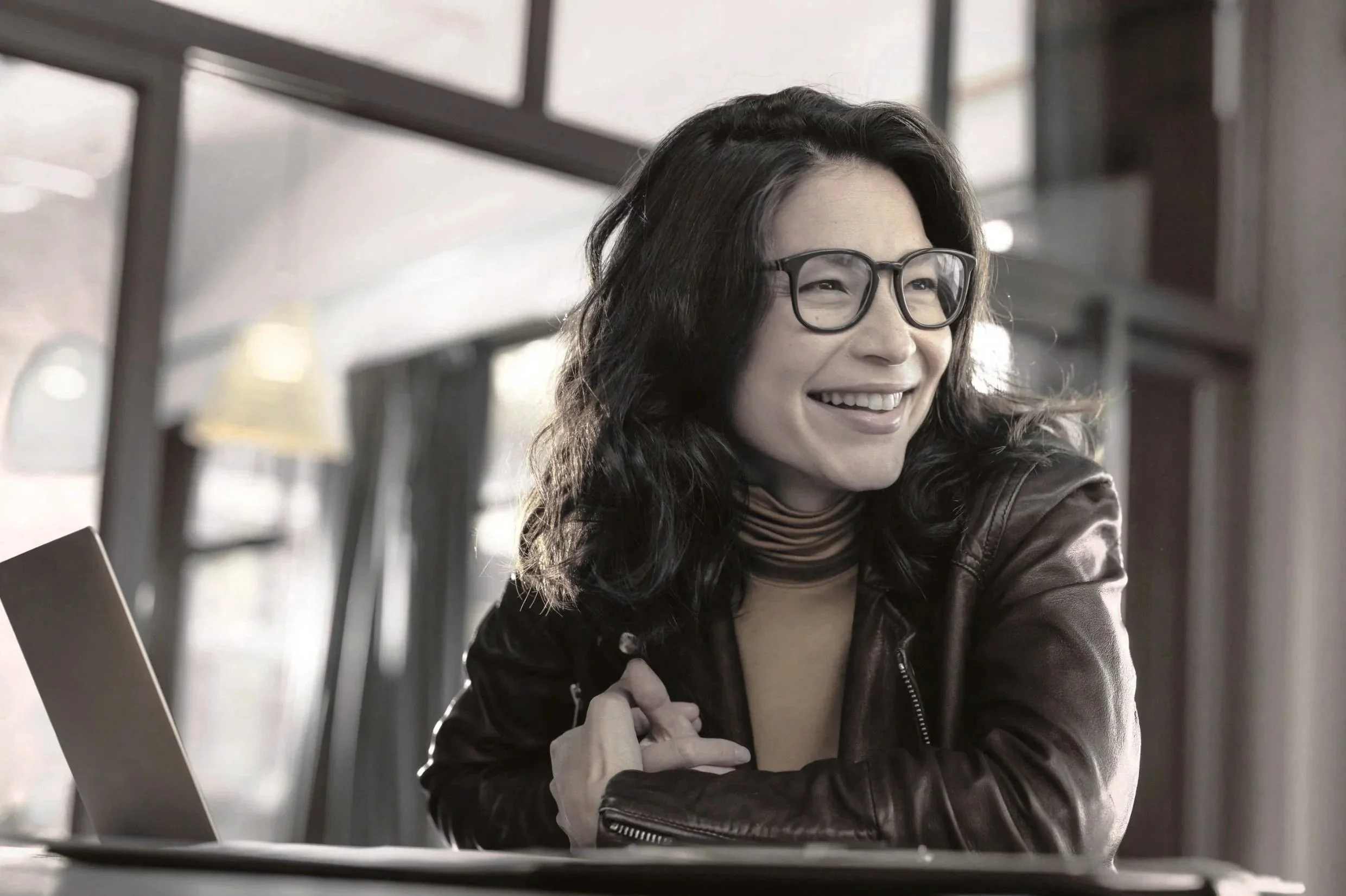 Lächelnde Frau mit schwarzen, lockigen Haaren, Brille und Lederjacke sitzt an einem Tisch in einem hell erleuchteten Raum, Foto canva