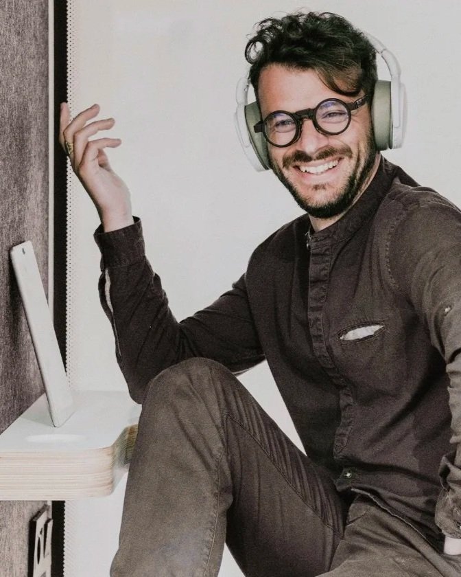 Mann mit Brille, Kopfhöhrer, dunkler Kleidung lächelt Bart und dunkle Kleidung