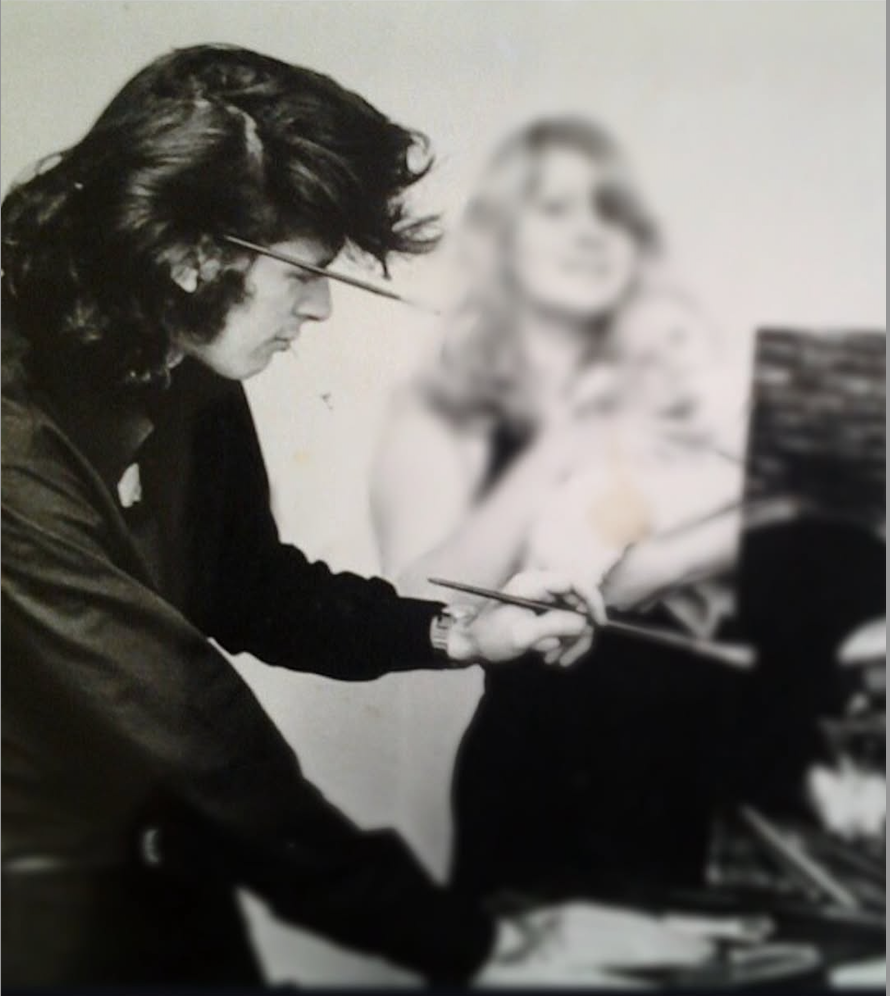 A man with long dark hair and glasses playing the piano with a woman in the background, out of focus