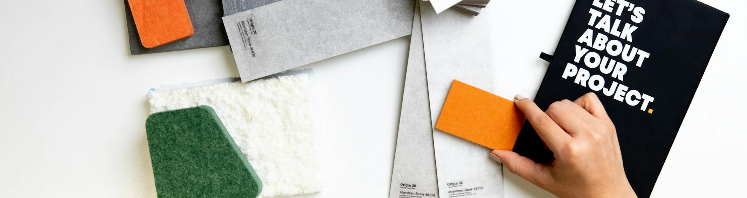 Ein Arbeitsplatz mit Papieren, Notizbüchern und einem Hand, die einen orangefarbenen Filzabschnitt auf einem Notizbuch platziert. Text auf einem Notizbuch lautet 'Let's talk about your project'.