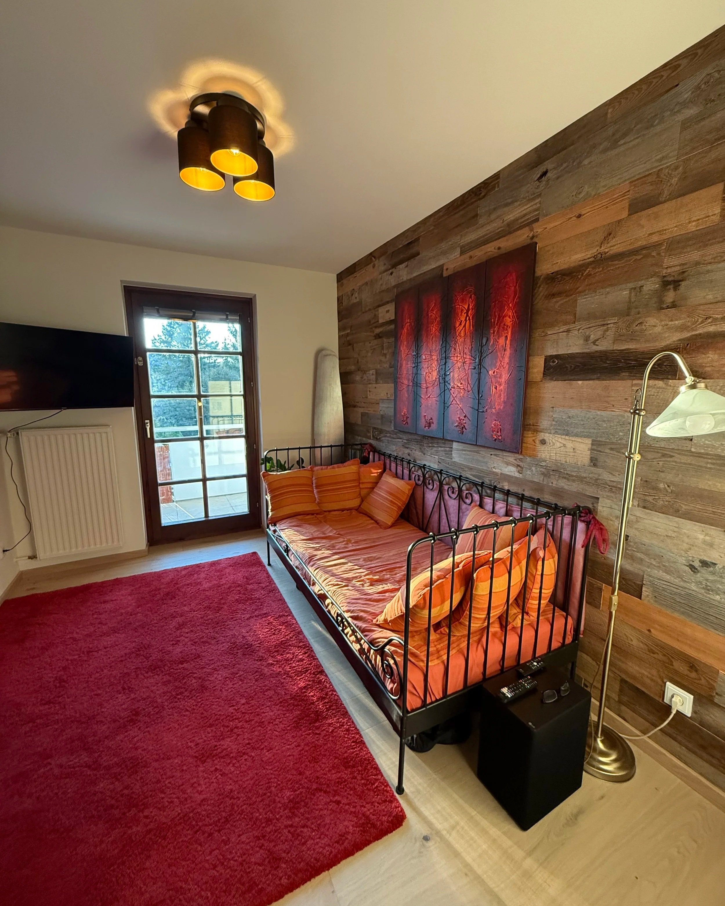 Warme Farben, ein Metaillgestellbett vor einer Altholzwand und Rottöne im Teppich sowie der dem Bild an der Wand