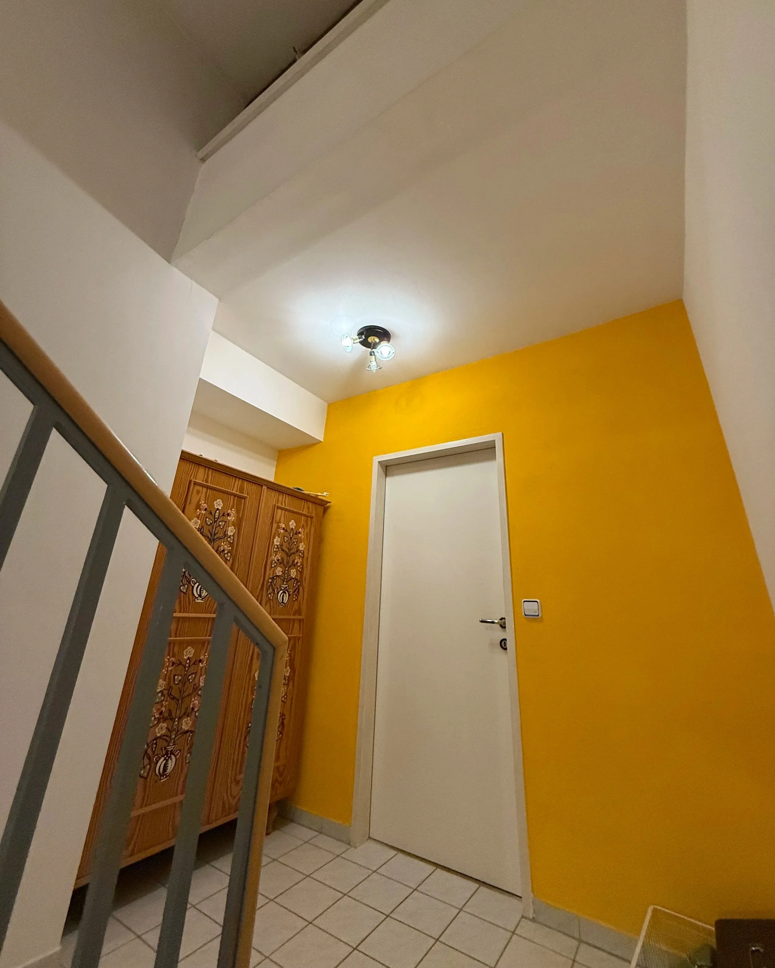 Das Stiegenhaus im Zwischengeschoss mit Blick zur Nische in der ein Bauernschrank steht. Die Wand rundherum um die Tür ist gelb. Weiße Fliesen.