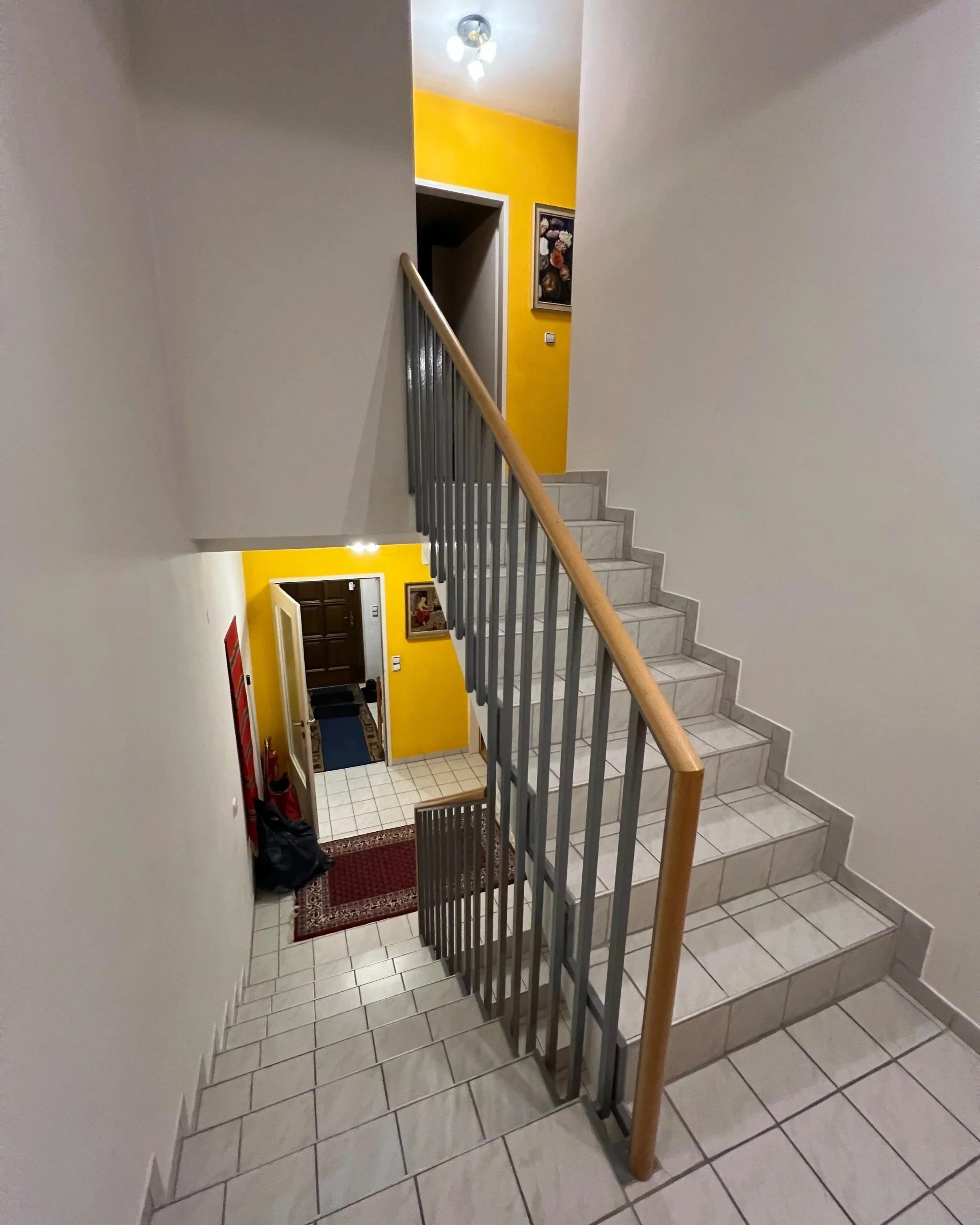 Stiegenhaus im Zwischengeschoss vor der Renovierung; weiße Fliesen, überwiegend weiße Wände, die Wandflächen auf der Ebene sind gelb