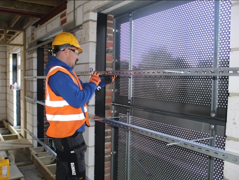 Construction worker in safety gear installing metal framing on perforated metal panels at a building site.