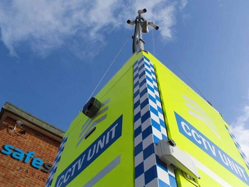 Close-up of a CCTV van with bright yellow and blue checkerboard pattern, mounted with security cameras, parked near a building with a sign that reads 'safes' against a partly cloudy sky.