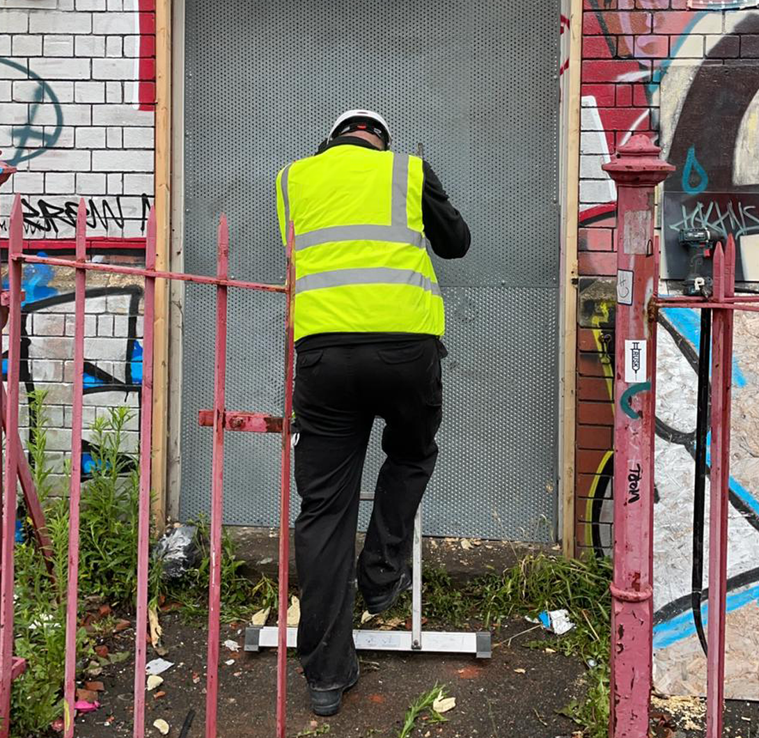A construction worker wearing a yellow safety vest and helmet working on a metal door or panel in front of a brick wall with graffiti. Pink metal fencing surrounds the area.