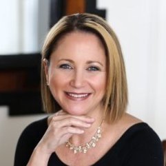 A smiling woman with shoulder-length blonde hair, wearing a black top and a pearl necklace, with her hand resting on her chin.