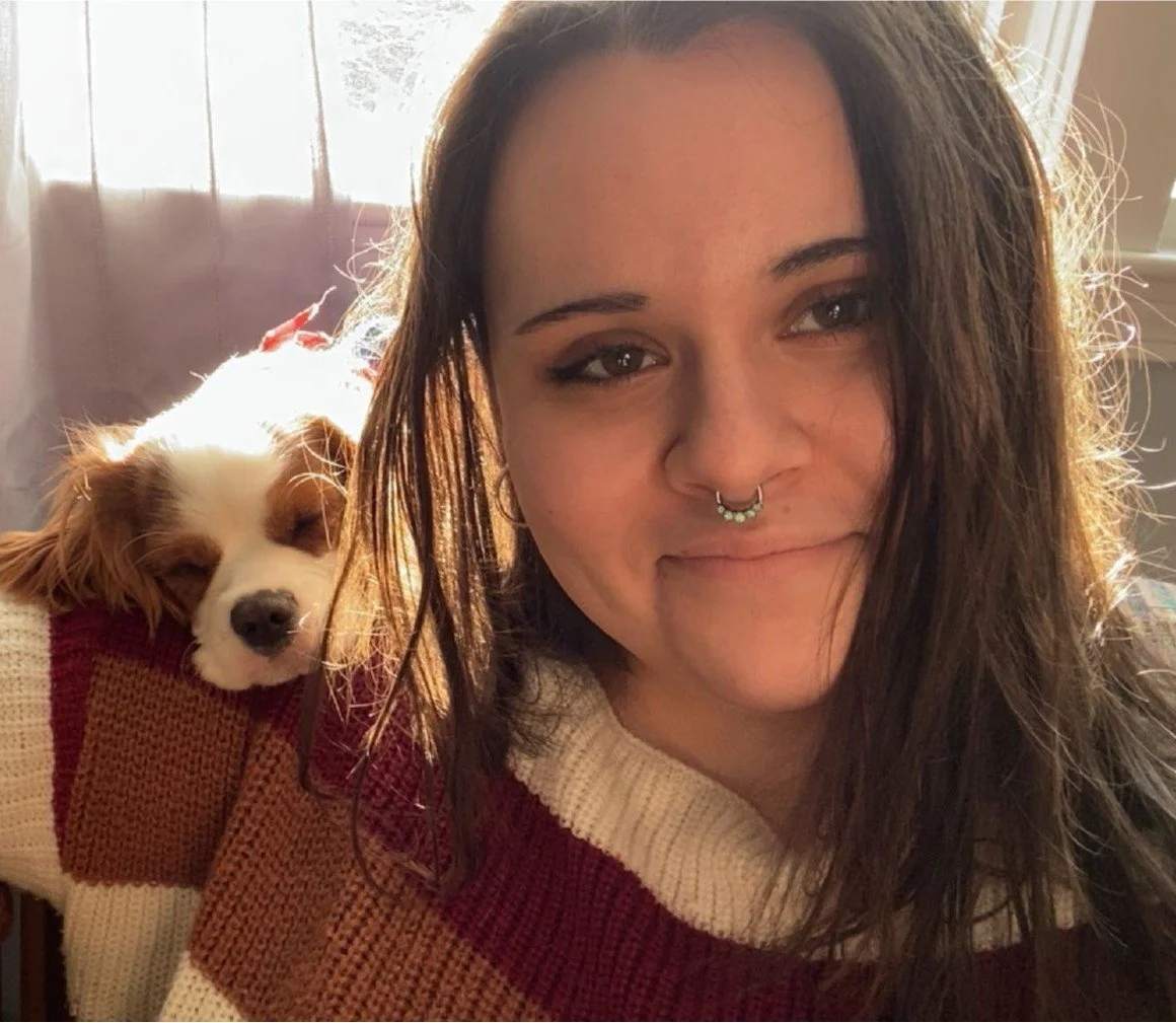 A young woman with brown hair, a septum piercing, and a smile, takes a selfie with a white and brown puppy resting on her shoulder in a sunlit room.