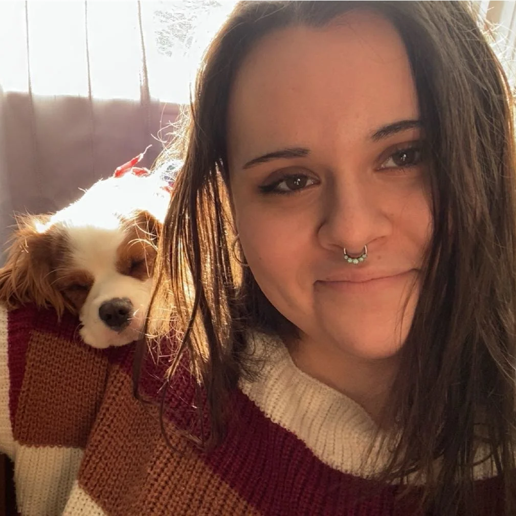 A woman with long brown hair, wearing a red and white sweater, smiling and sitting with a small, white and brown dog that's resting its head on her shoulder.