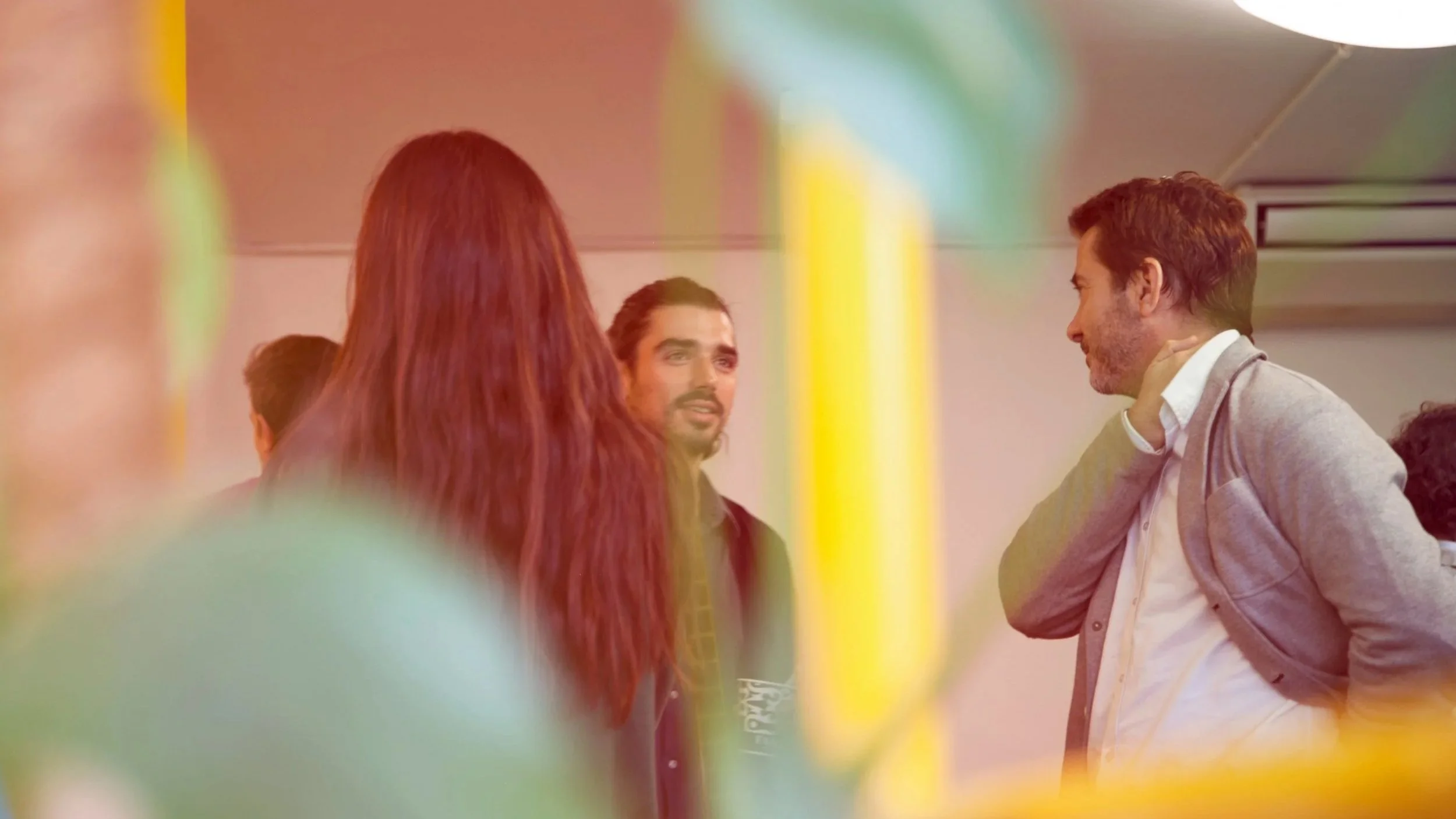 A group of people engaged in conversation indoors, with a man in a gray blazer and white shirt smiling and holding his neck, a woman with long red hair, and other individuals with colorful lighting reflections around.