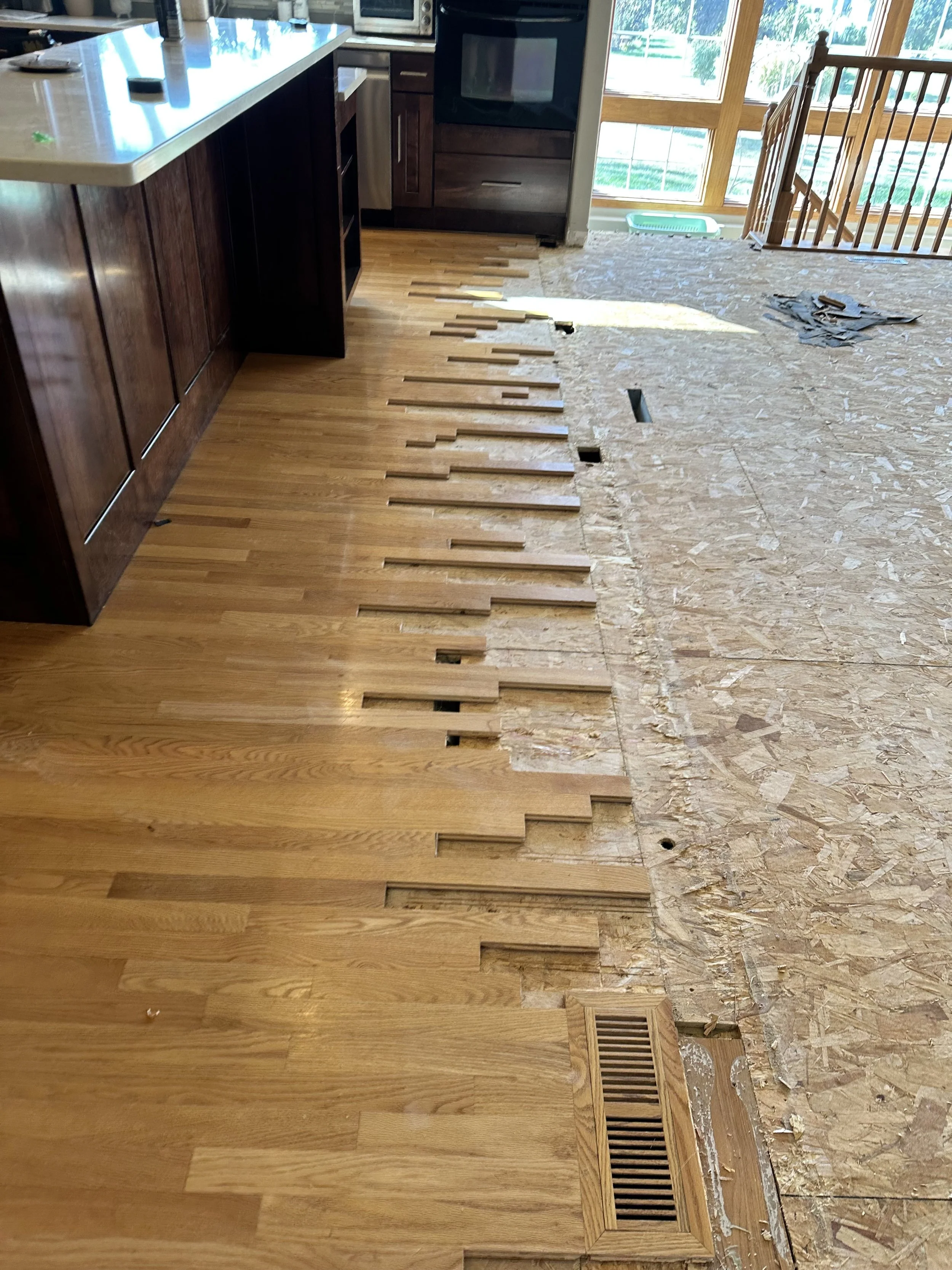 Kitchen hardwood flooring being replaced, with partially removed wooden floorboards revealing subflooring underneath.
