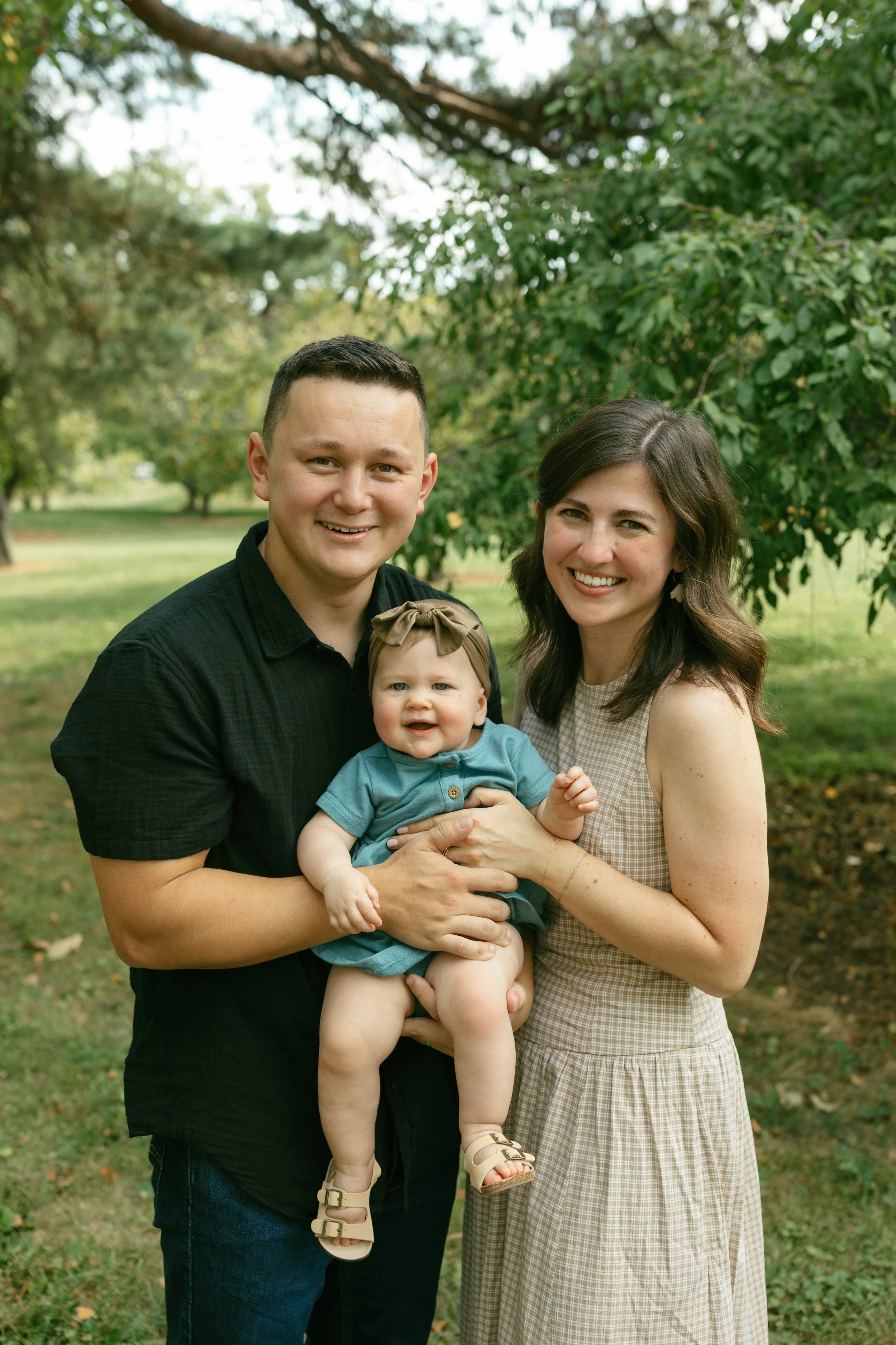 A happy family of three standing outdoors in a park with trees in the background. A man and woman are smiling while holding a smiling baby girl between them. The man wears a black shirt, the woman a sleeveless beige dress, and the baby has a bow headband and sandals.
