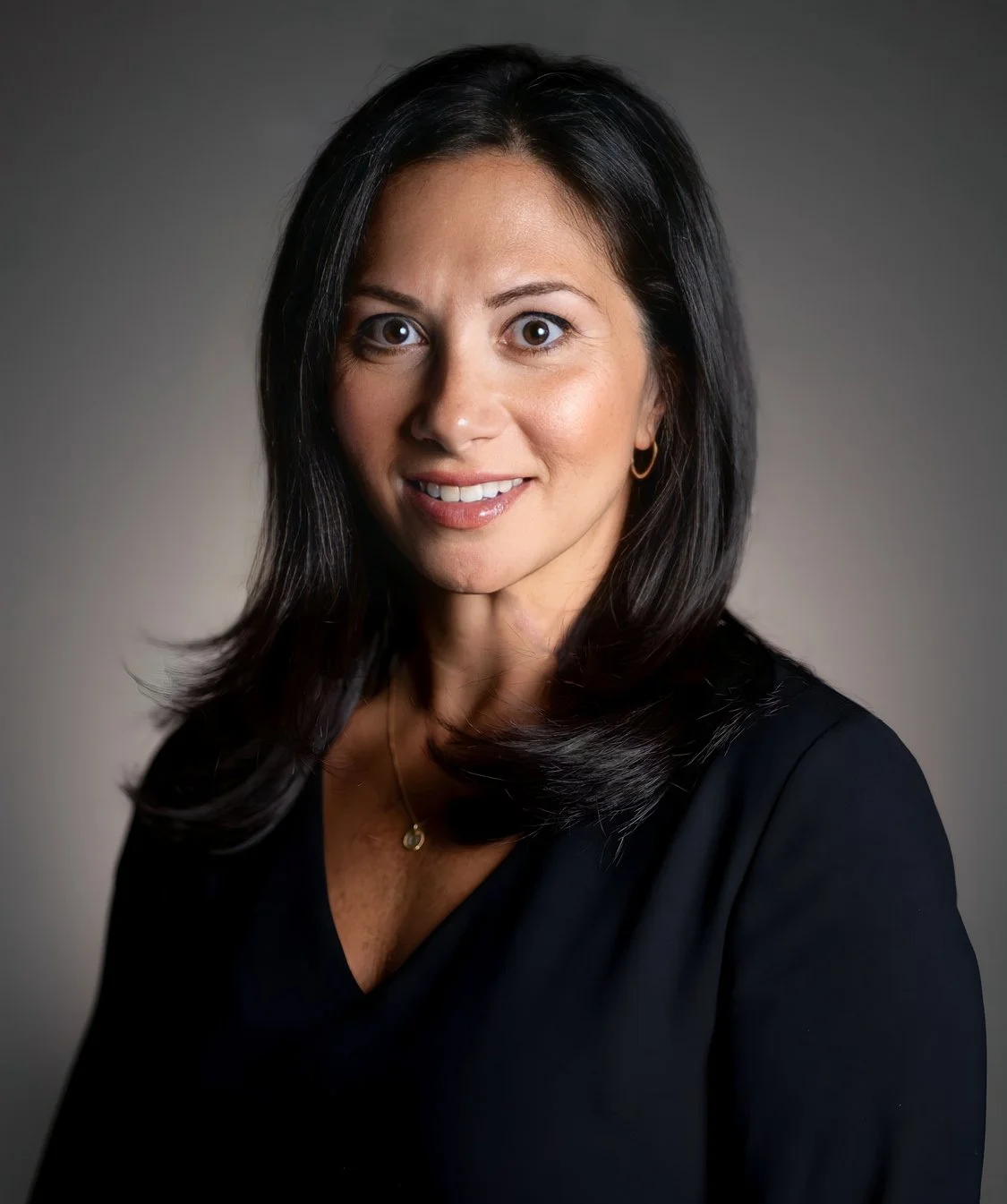 A woman with shoulder-length black hair, wearing a black top and gold jewelry, smiling softly against a neutral background.