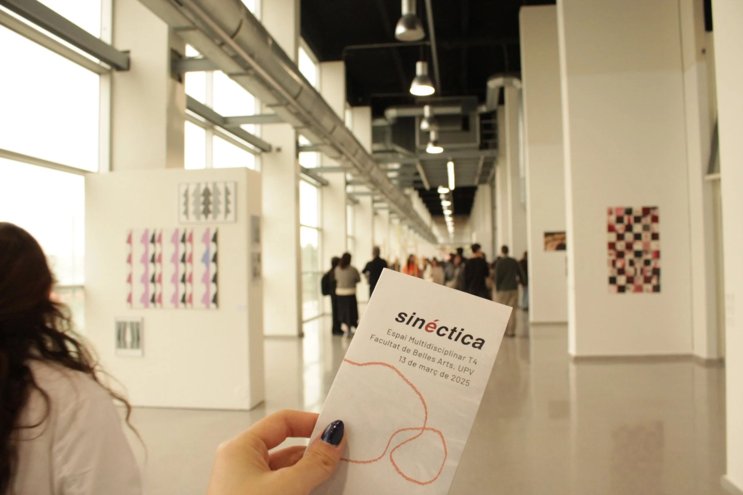 Persona sosteniendo un folleto en una galería de arte llena de obras y visitantes