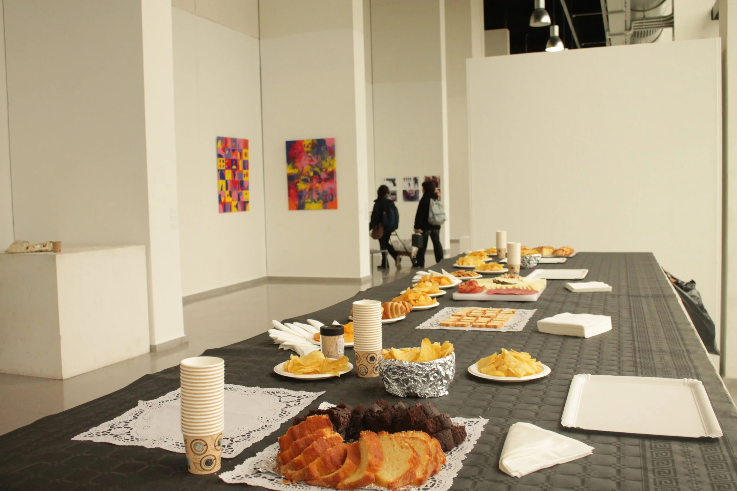Mesa de buffet con pasteles, galletas, papas fritas y bebidas en una galería de arte con cuadros en las paredes.