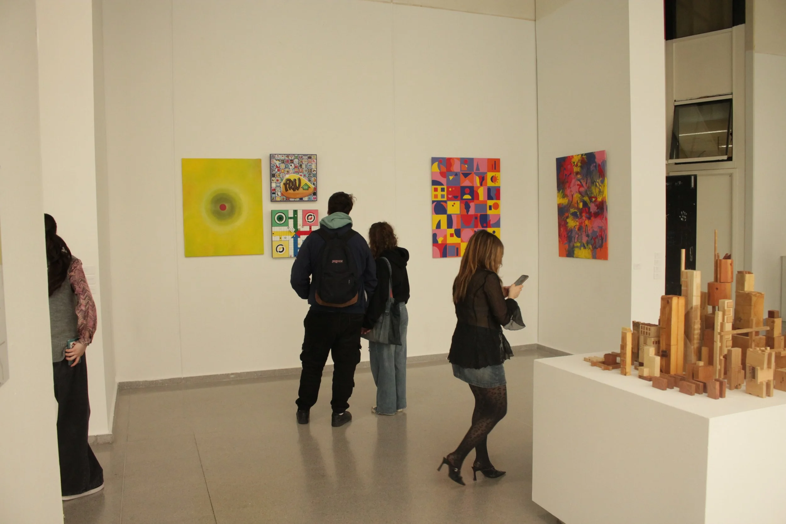 Personas observando obras de arte en una galería, con cuadros abstractos y decoraciones en las paredes, y esculturas de madera en exhibición.