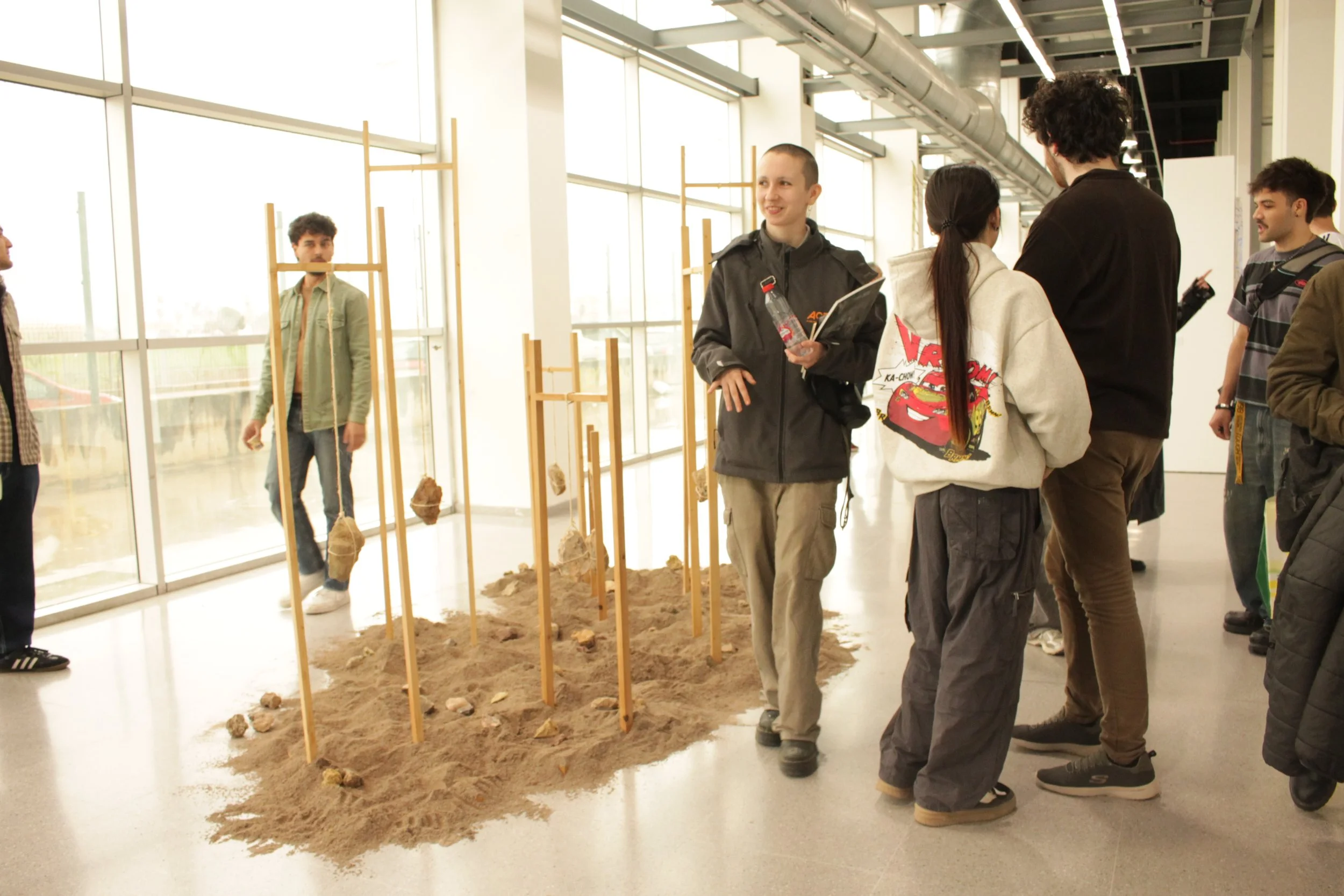Grupo de adolescentes conversando en una exhibición en un espacio interior con estructura metálica y ventanas grandes, junto a una zona de arena con columnas de madera y piedras.