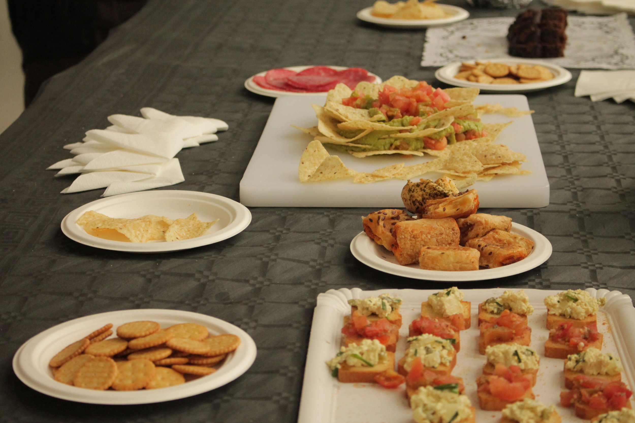 Variedad de alimentos en platos desechables en una mesa, incluyendo tacos con guacamole y tomates, galletas saladas, empanadas, y pequeños bocados con ingredientes diversos.