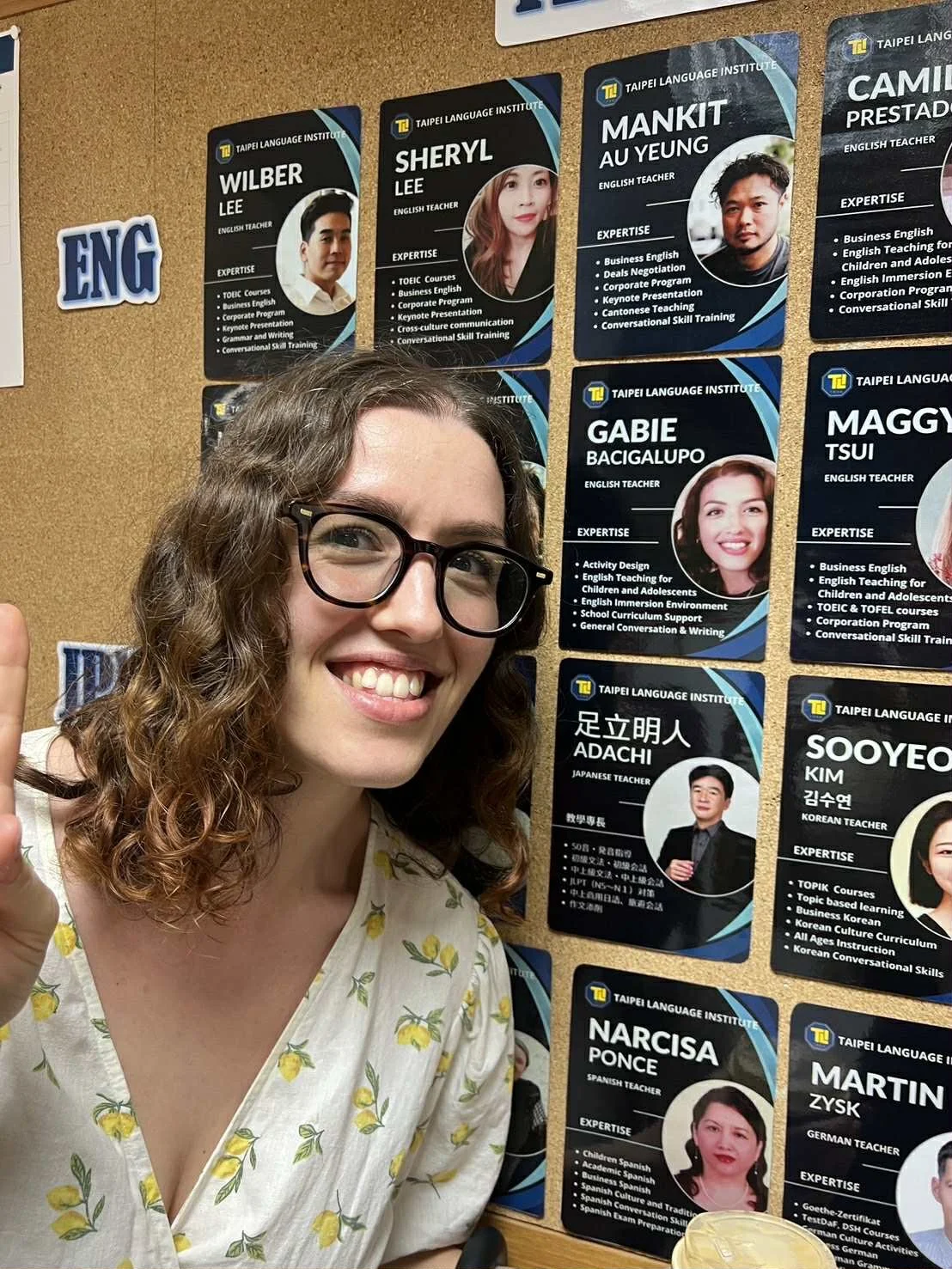 A woman with curly hair and glasses smiling and pointing at a wall of teacher profiles at Taipei Language Institute, featuring various language teachers with their expertise and credentials.