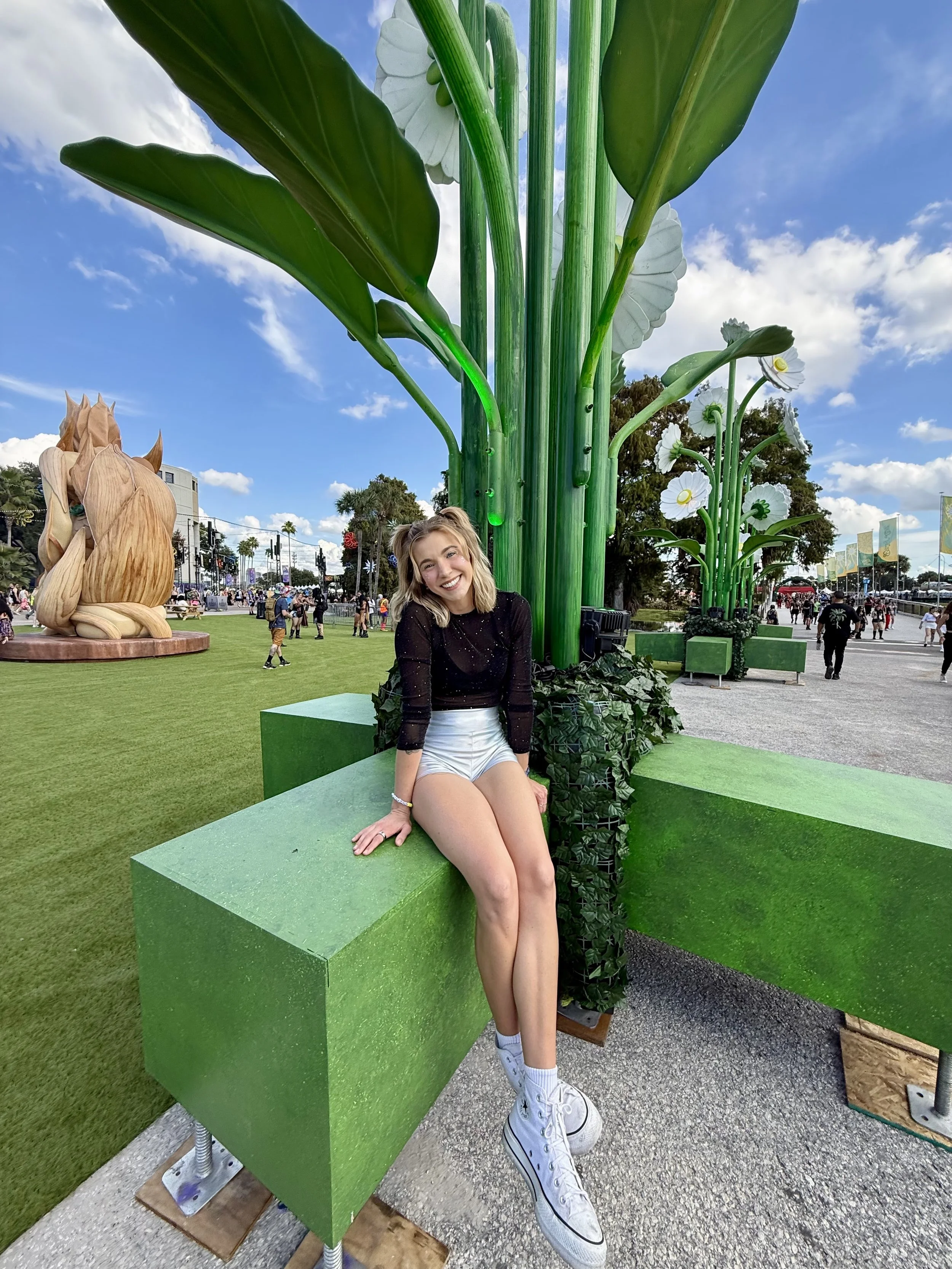A young woman with blonde hair sitting on a green bench at an outdoor event, smiling. Behind her are tall, oversized green plant sculptures with large leaves and white flowers, set against a partly cloudy sky. There are people walking in the background, and a large lion sculpture nearby.