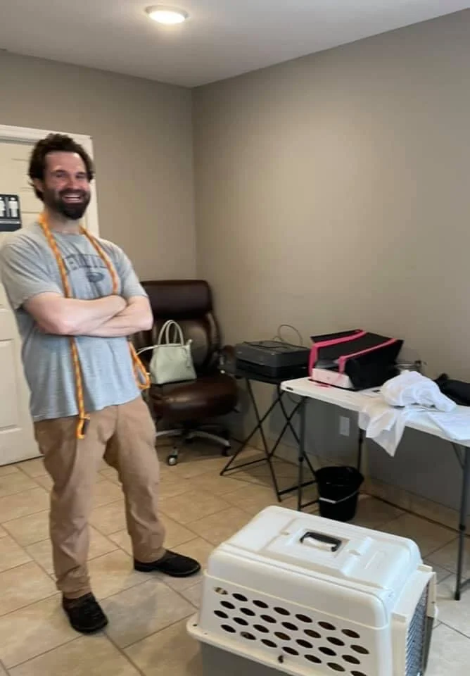 A man standing in a room with a smile, crossing his arms. He has a measuring tape around his neck. The room has a chair, a desk with papers and office supplies, a printer, and a pet carrier on the floor.