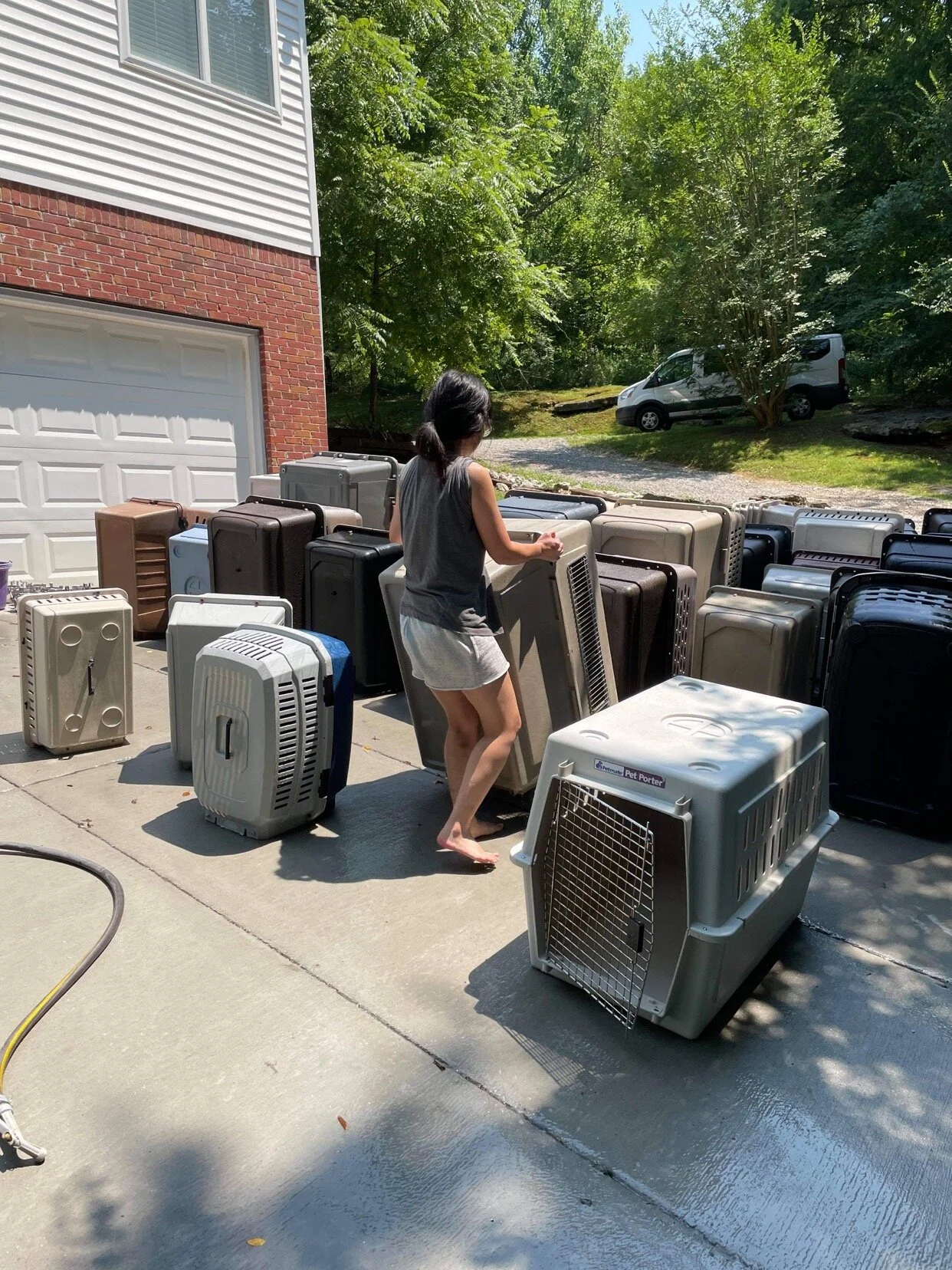 A woman is outdoors on a concrete driveway, surrounded by numerous pet carriers. She is bending over and holding one of the carriers, with a residential garage and trees in the background.