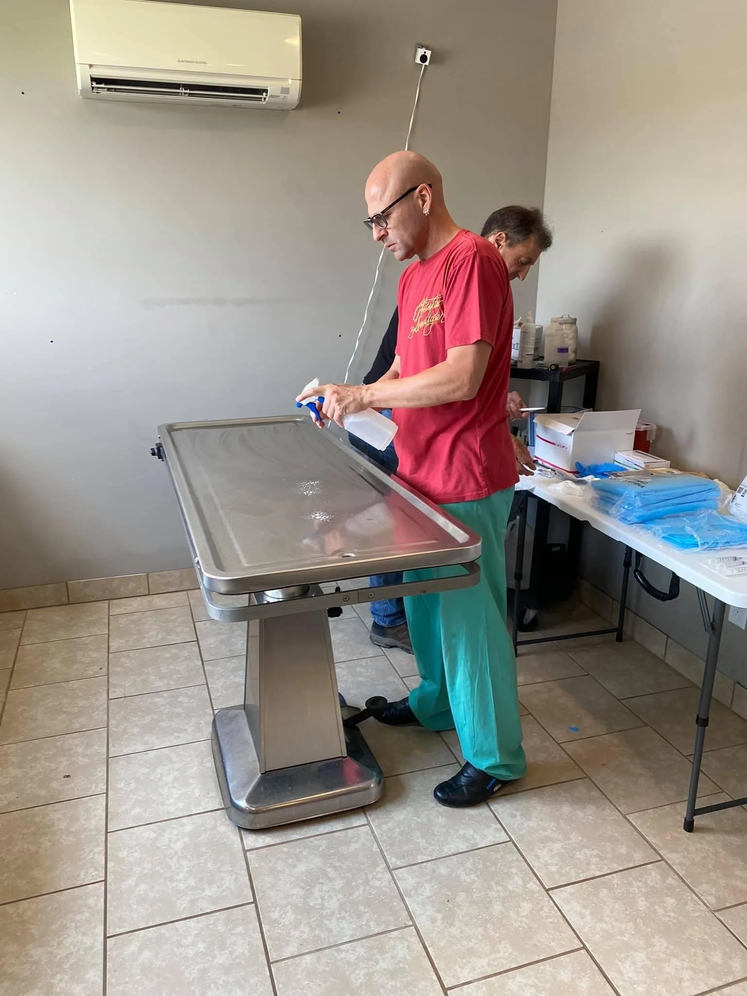 A man wearing glasses, a red shirt, and green medical scrubs, standing at a stainless steel examination table in a medical setting, holding a spray bottle. Two other individuals are working at a table in the background, with medical supplies and equi