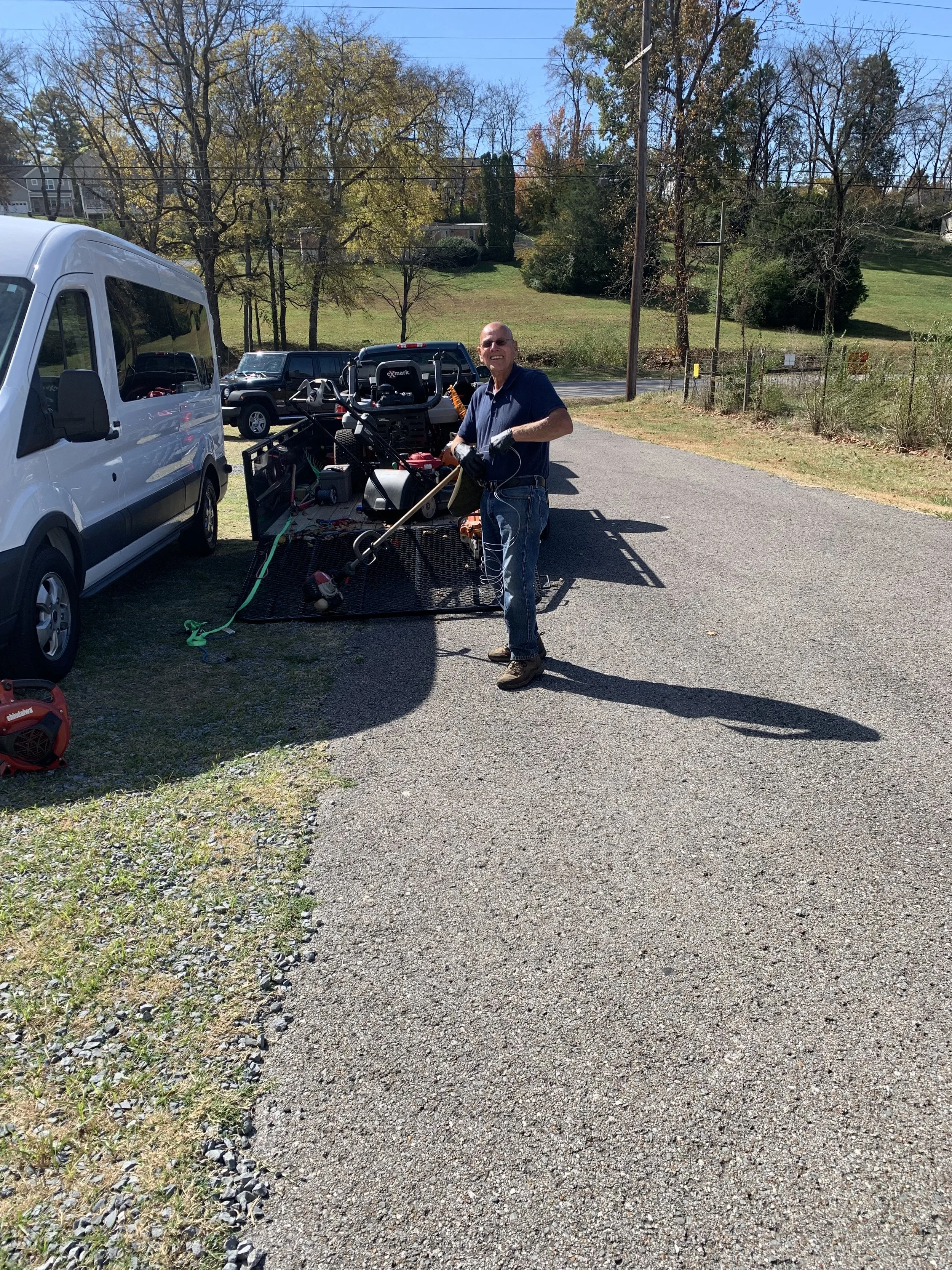 A man standing outside in a parking lot, holding a weed trimmer, with a trailer full of yard tools and equipment behind him. There are trees and grass in the background with clear blue skies.