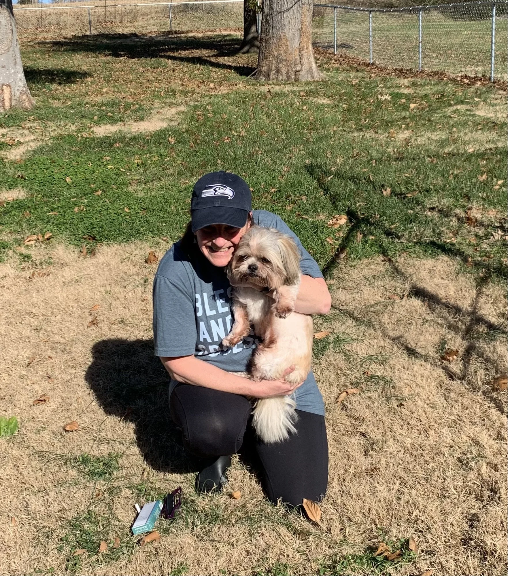 A woman kneeling on grass and holding a small dog outdoors on a sunny day.
