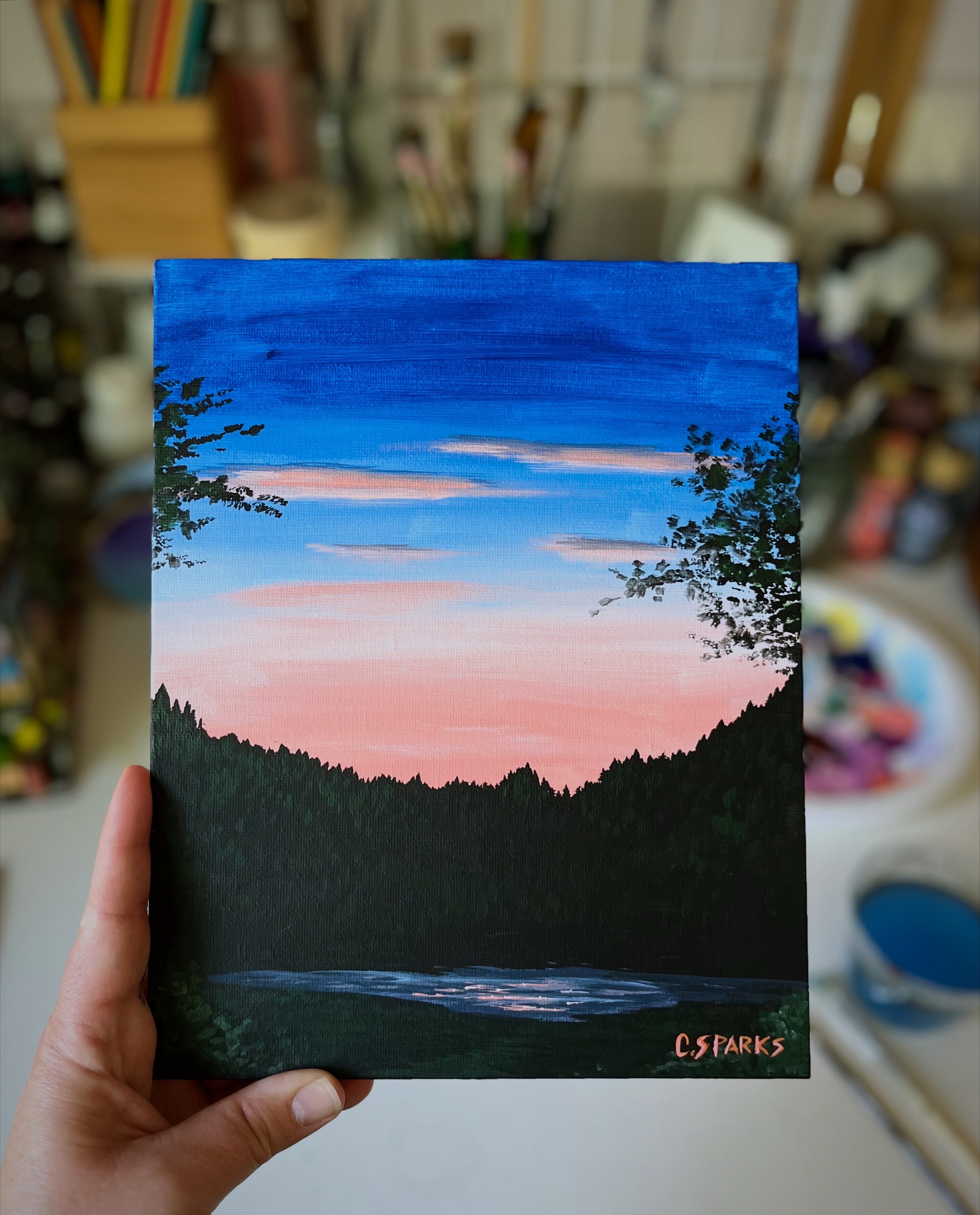 a painting of a blue and peachy pink sky above a forest and lake. The lake is reflecting glimmers of light as the sun appears to be rising behind the dark green and black forest silhouette.