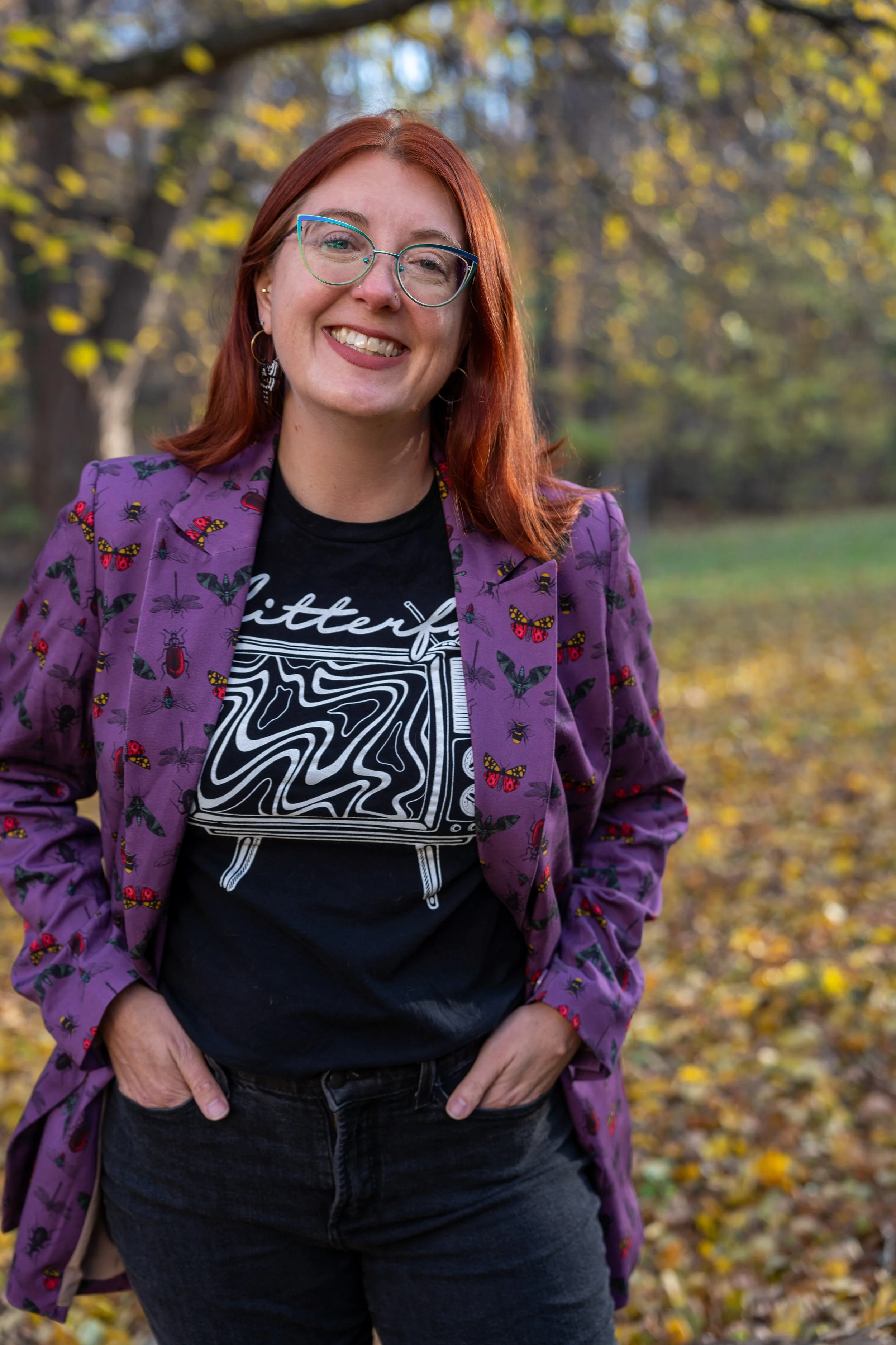 A photo of Brigitte. She is white and has red hair and glasses. She smiles, wearing a black graphic t-shirt, purple jacket with butterfly and insect print, and black jeans. Leaves are on the ground and trees in the background.