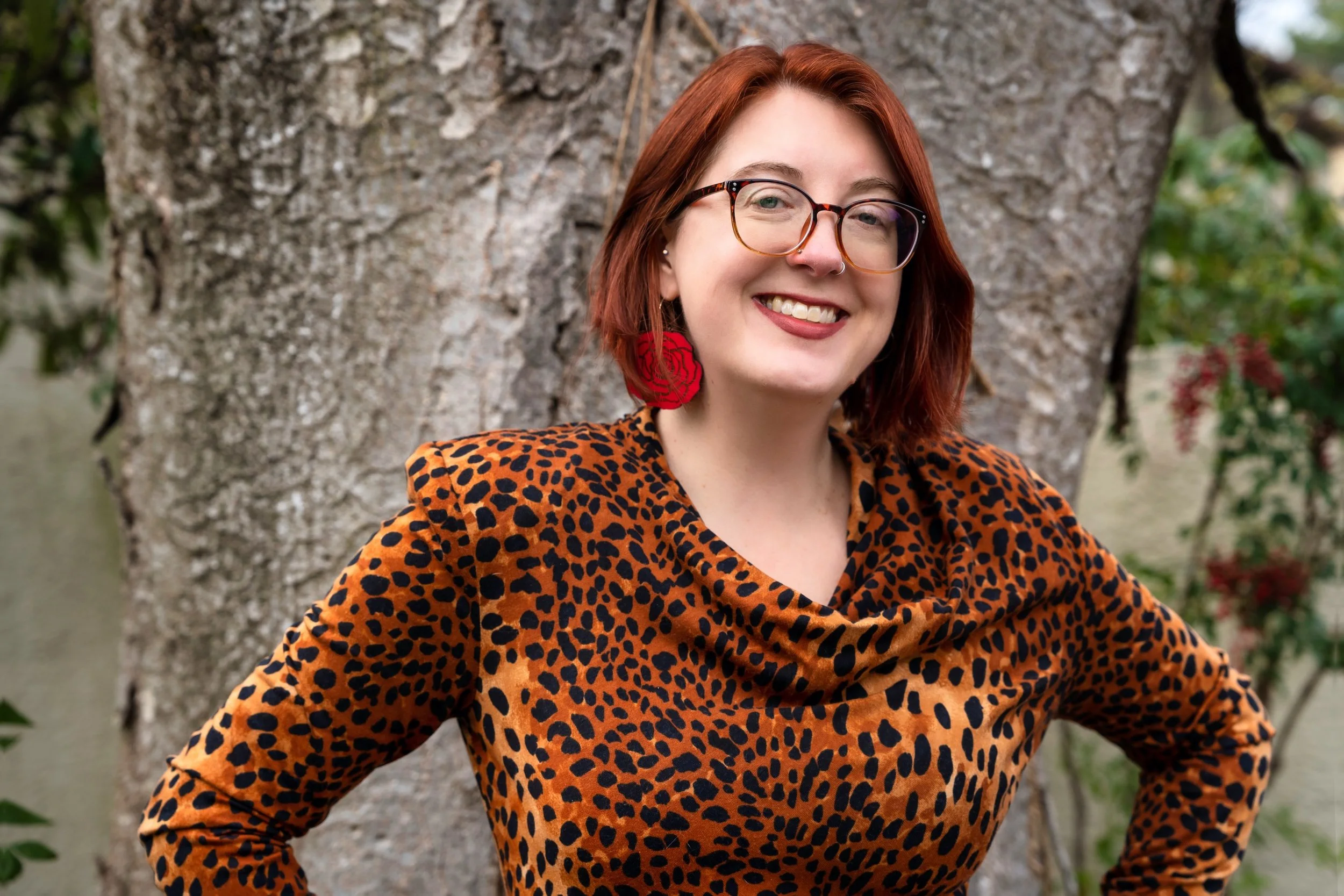 A smiling woman with short red hair, glasses, and a nose ring, standing outdoors in front of a tree, wearing a leopard print top and large red rose earrings.