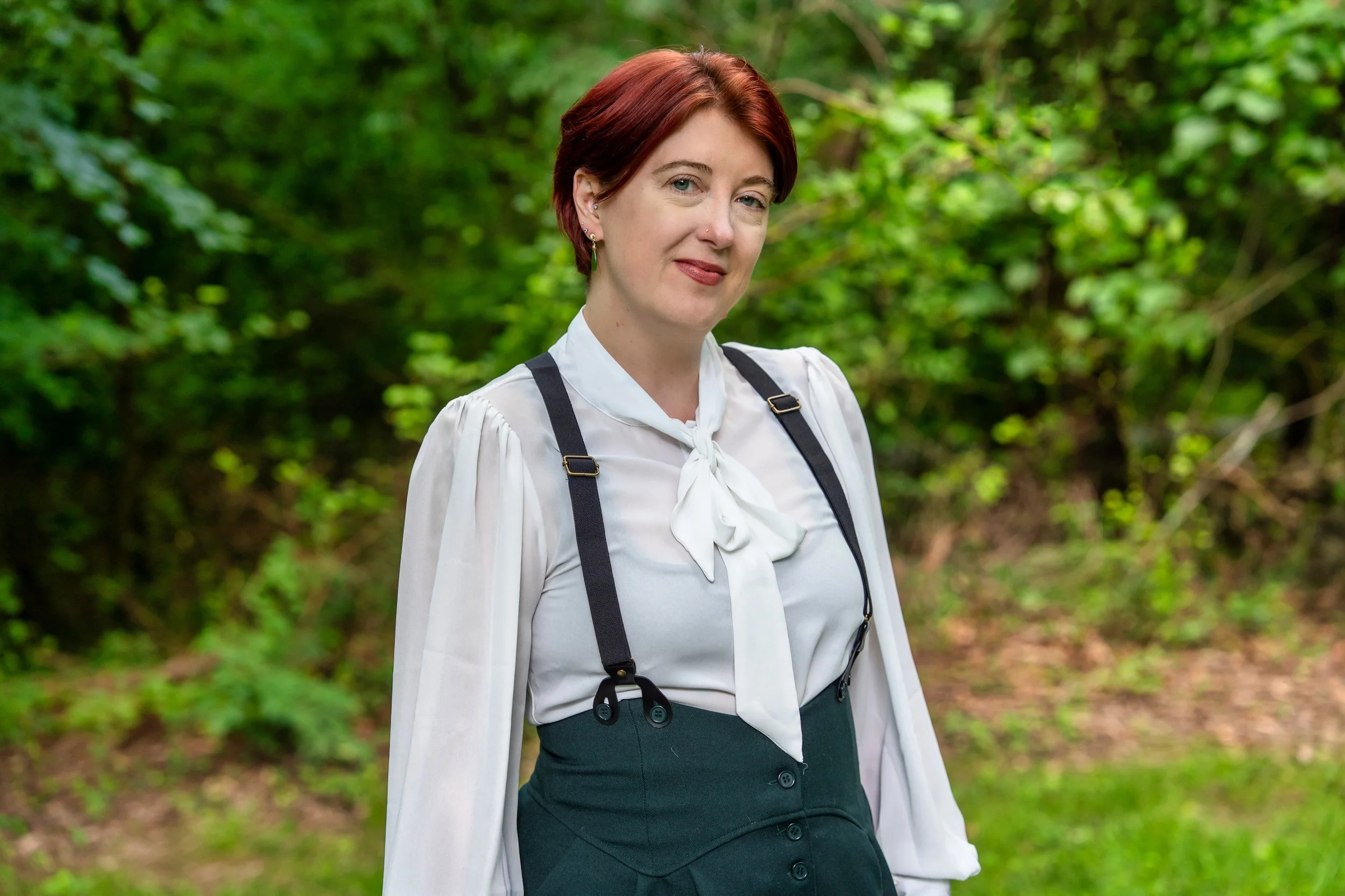 A person with short red hair, wearing a white blouse with a bow, dark skirt, and suspenders, standing outdoors in a green wooded area.