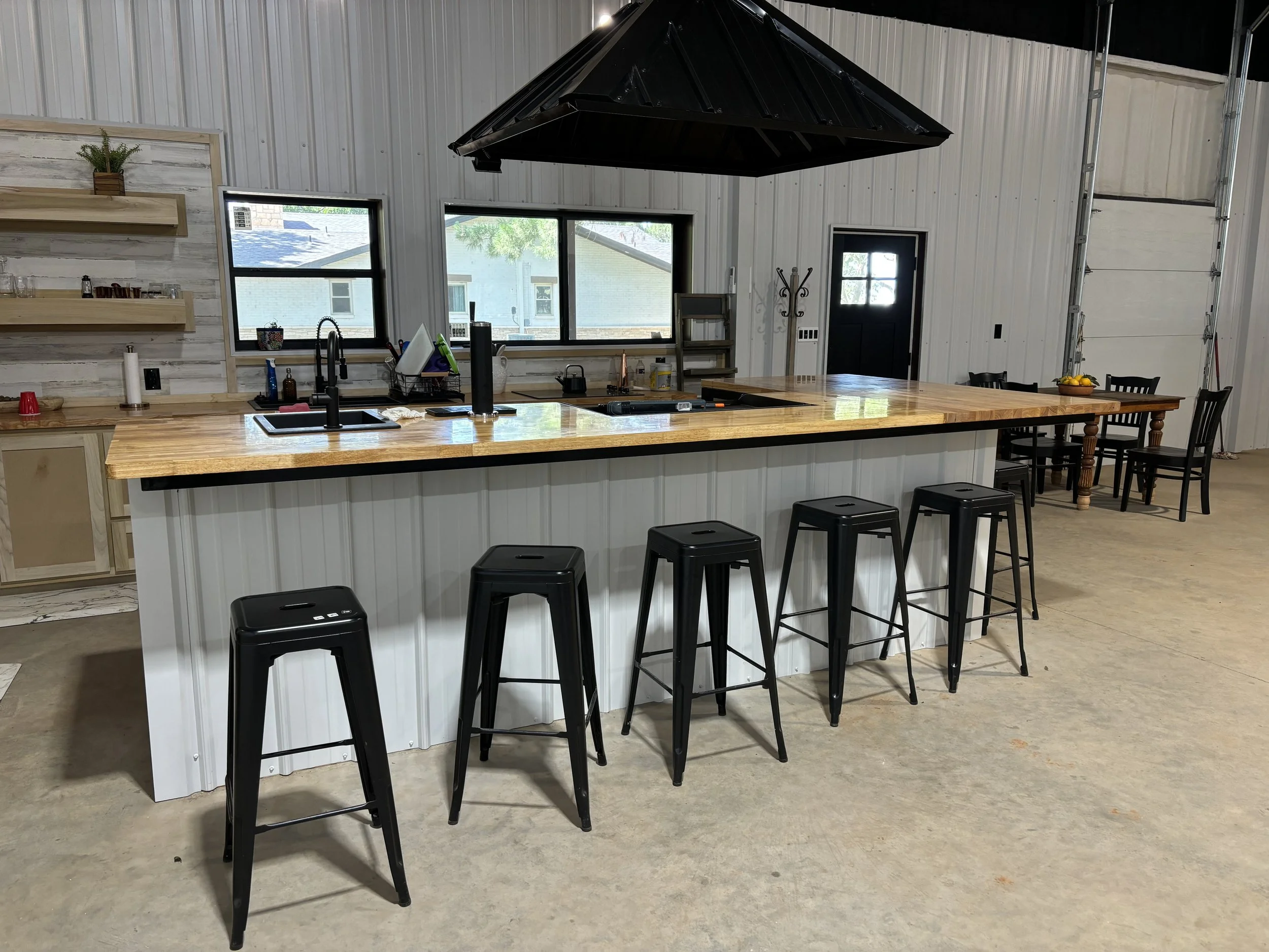 A modern kitchen with a large wooden island counter, black bar stools, white shiplap walls, windows showing a neighborhood outside, and a black range hood above.