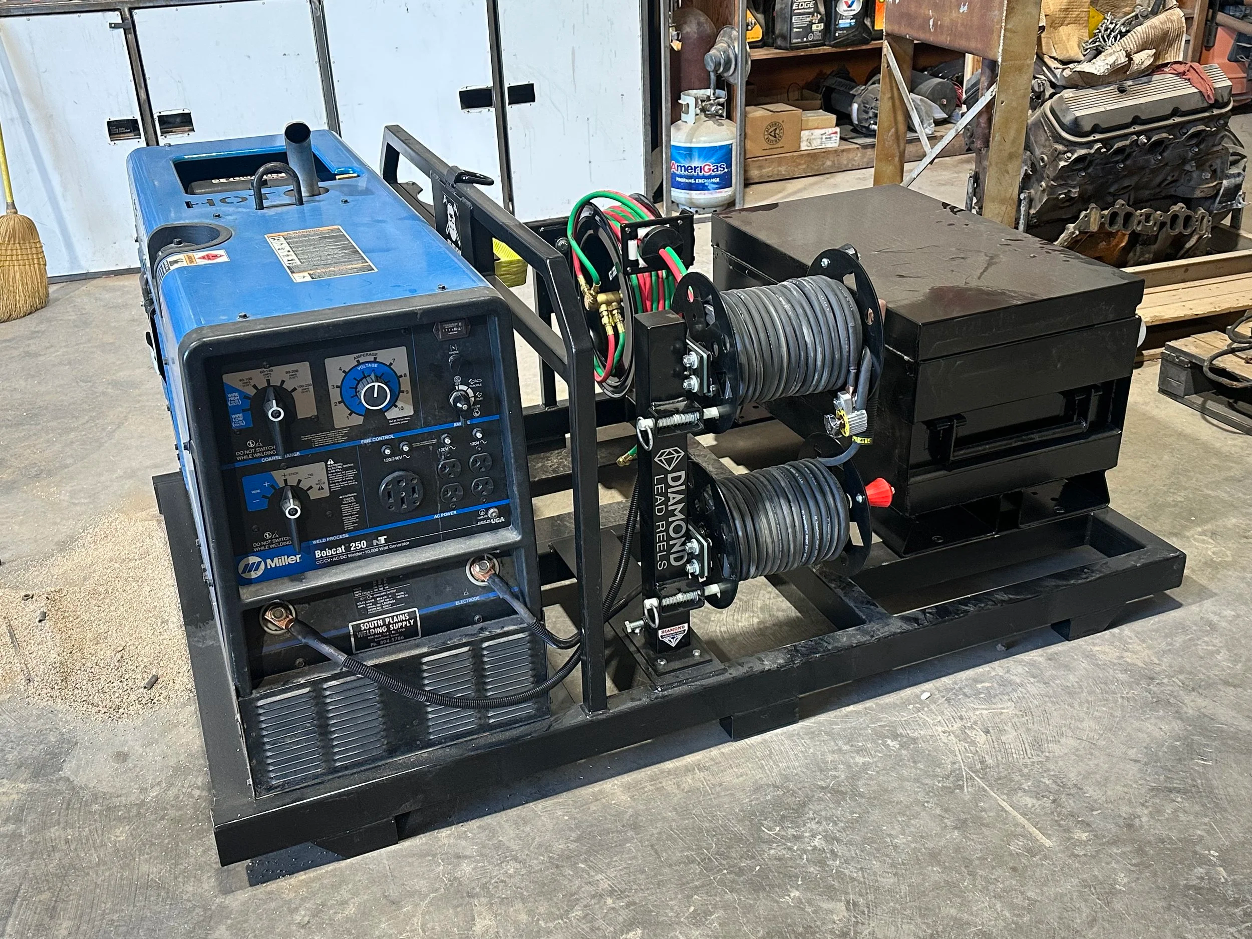 A mobile welding setup featuring a blue Miller Bobcat 250 NT generator on a black metal cart. The setup includes a reel of welding cable with color-coded connections, various controls and a display on the generator, and is situated in a garage or workshop environment with tools, engine parts, and a propane tank visible in the background.