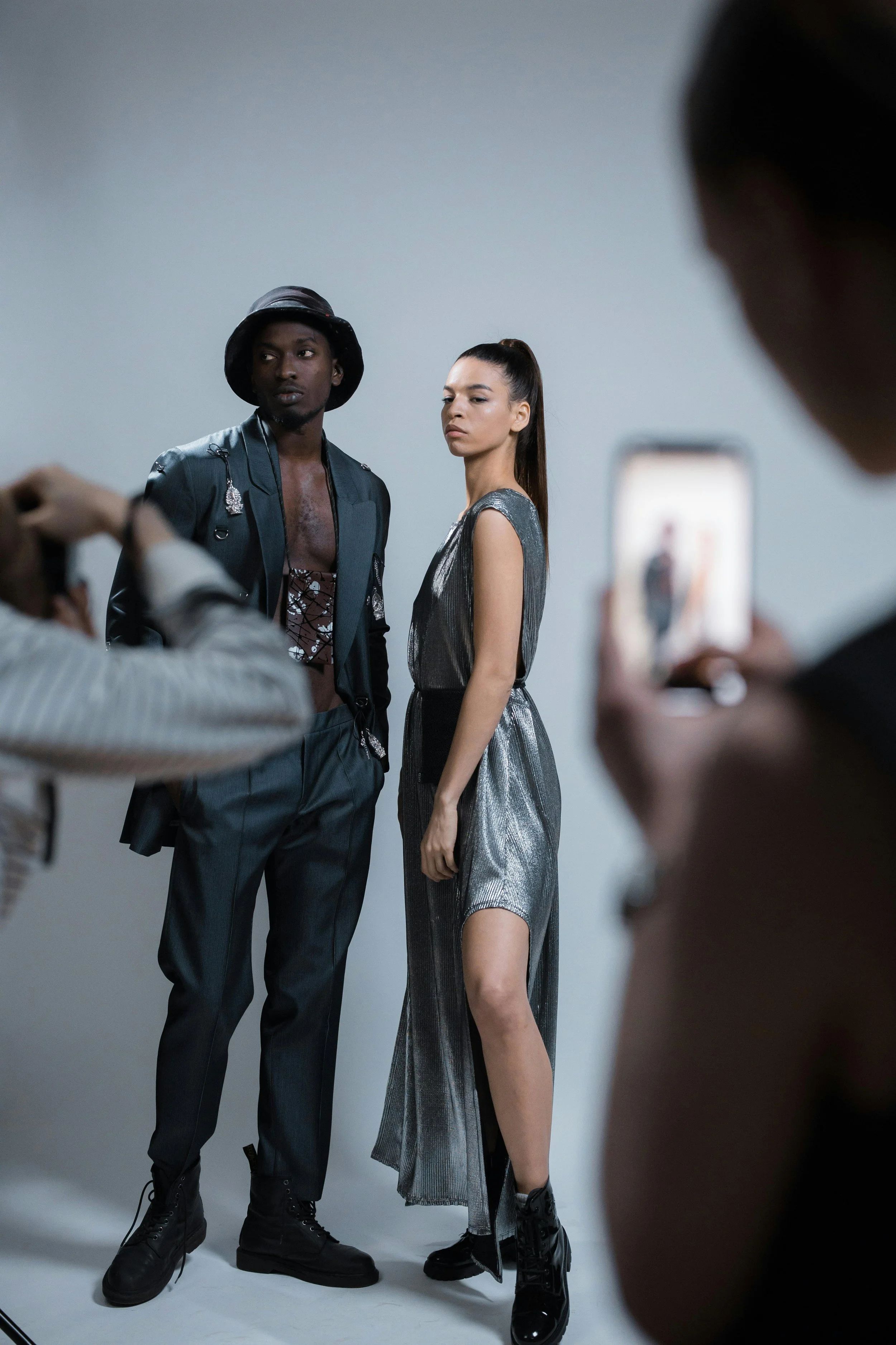 Two models pose for a photoshoot against a gray background, while a person takes a picture of them with a smartphone. The male model is wearing dark pants, a bucket hat, and a metallic jacket, while the female model is in a metallic dress with a high slit.