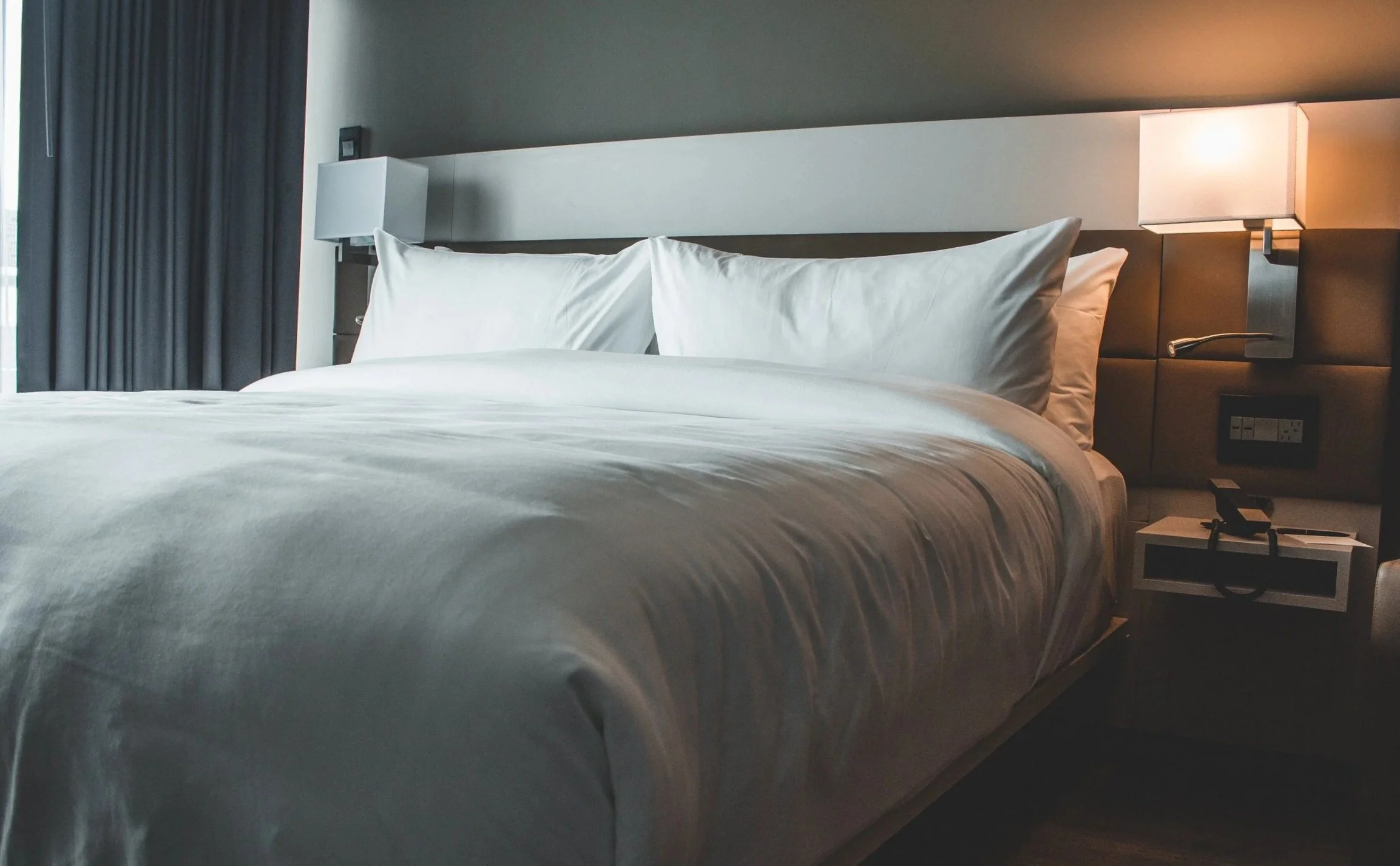 A neatly made hotel bed with white linens and pillows, flanked by modern wall-mounted lamps in a contemporary hotel room.