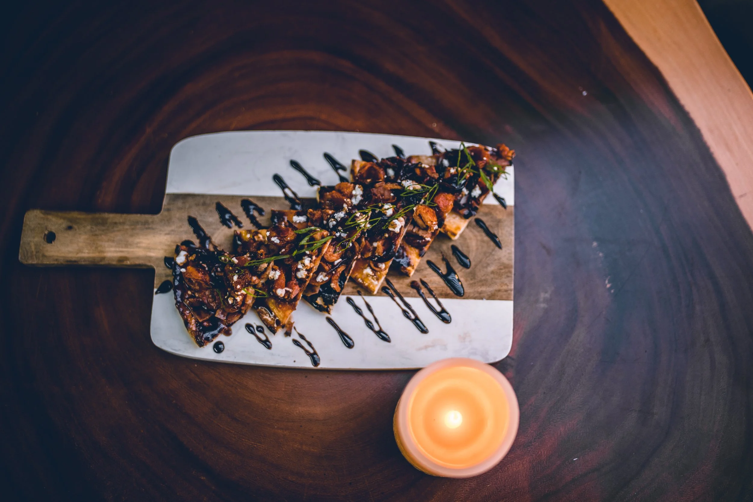 A slice of pizza topped with chicken, herbs, and drizzled with balsamic glaze, served on a cutting board with a candle nearby.